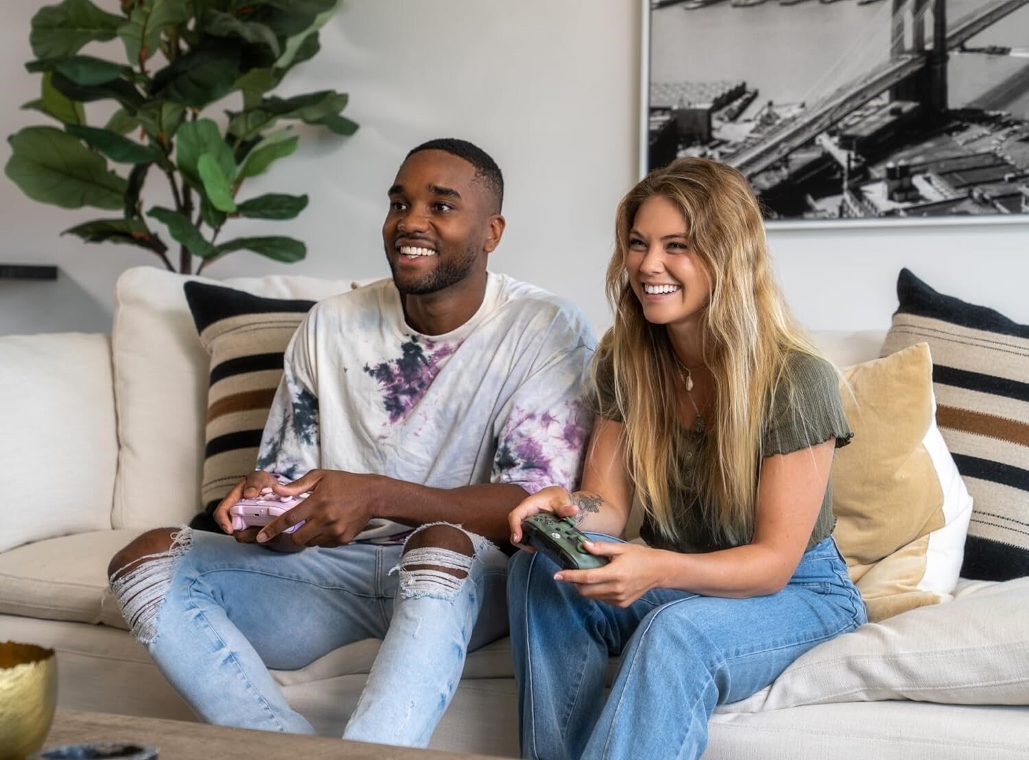 two people sit on a couch while holding gaming controllers and smiling