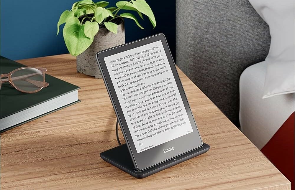 the kindle paperwhite signature edition sits on a wireless charger on a nightstand.