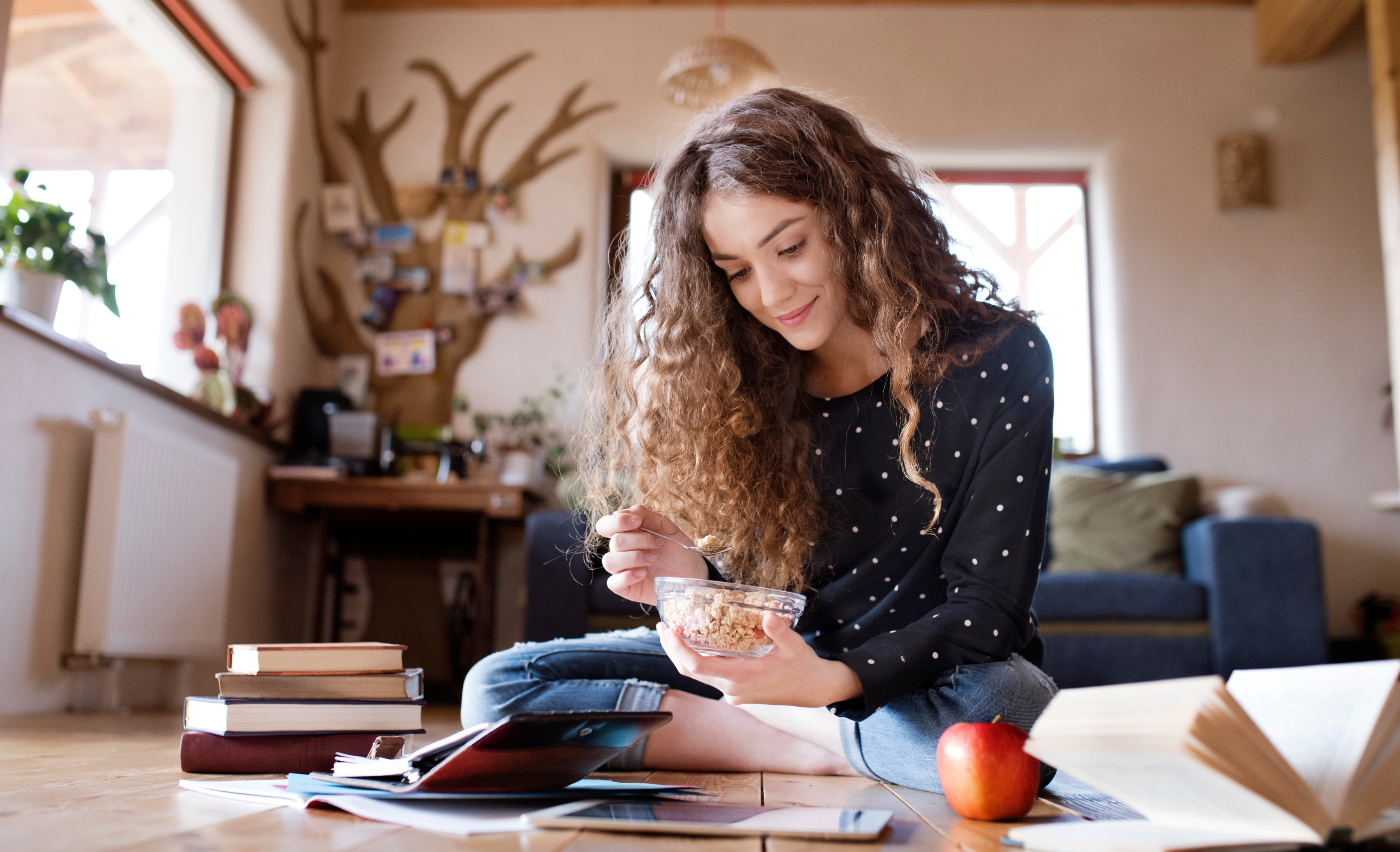 Female student sitting on floor, reading books, studying for final exam at university. Beautiful student at home eating cereals.