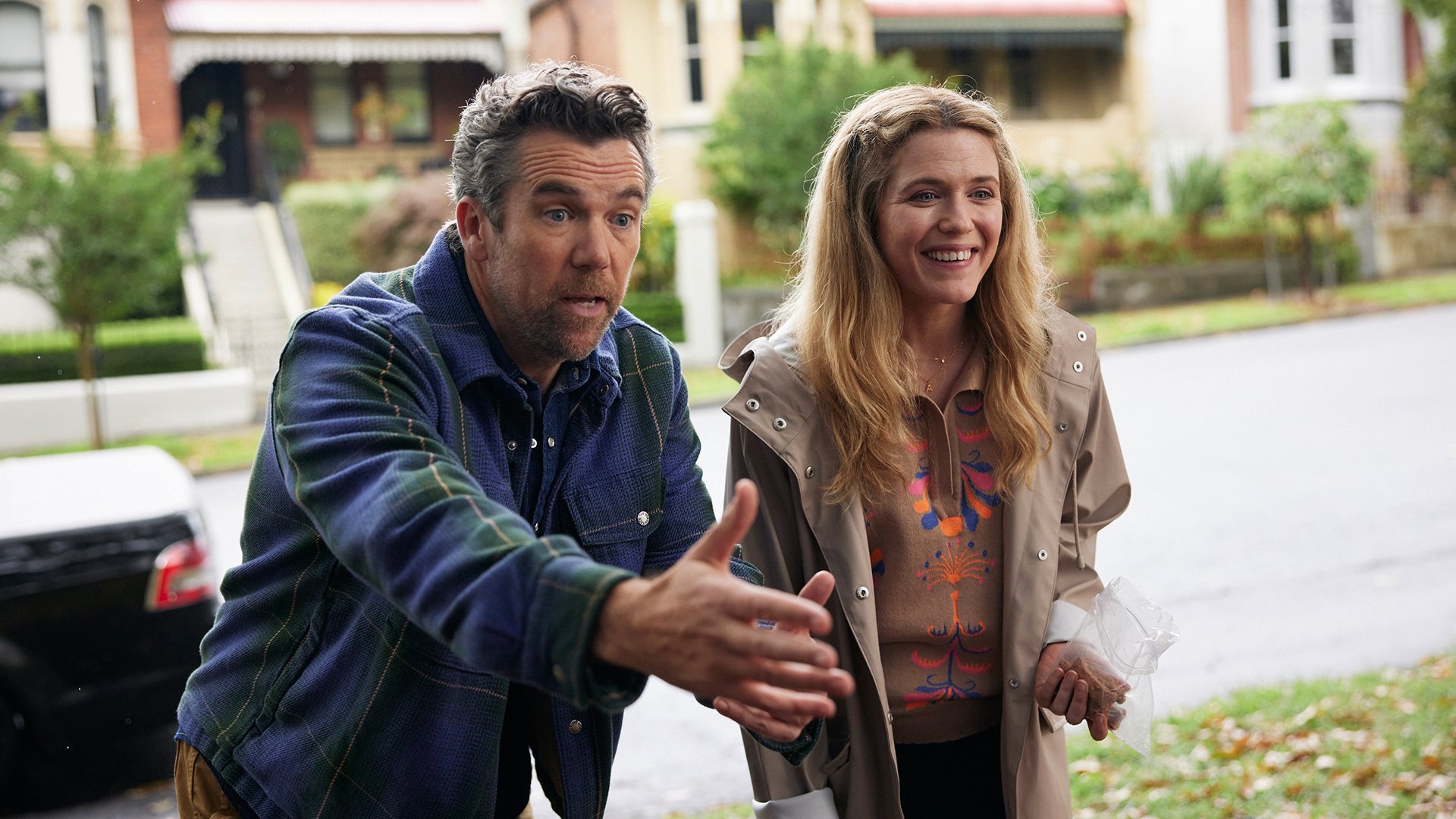 A man and a woman stand on the doorstep of a suburban road, looking excited.