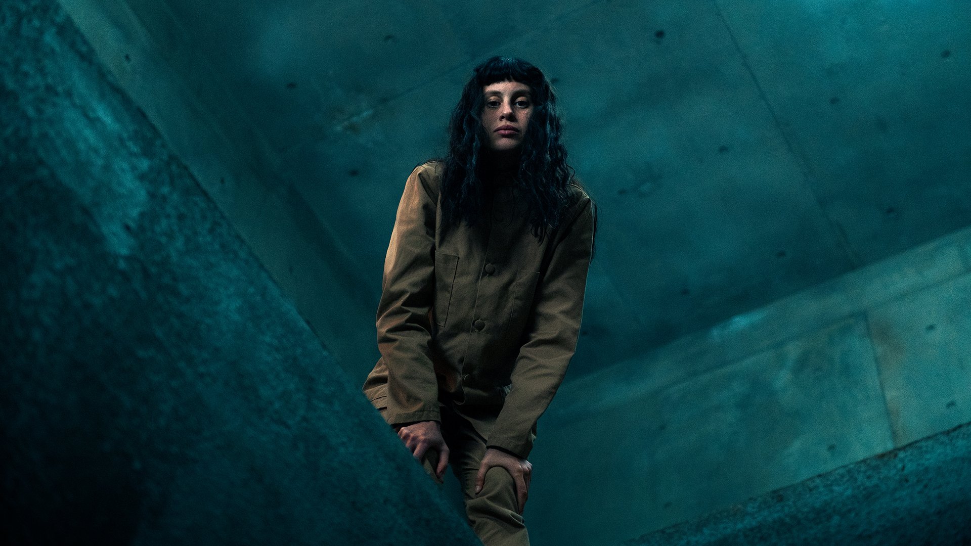 A woman stands on the edge of a concrete platform, peering down towards the camera.