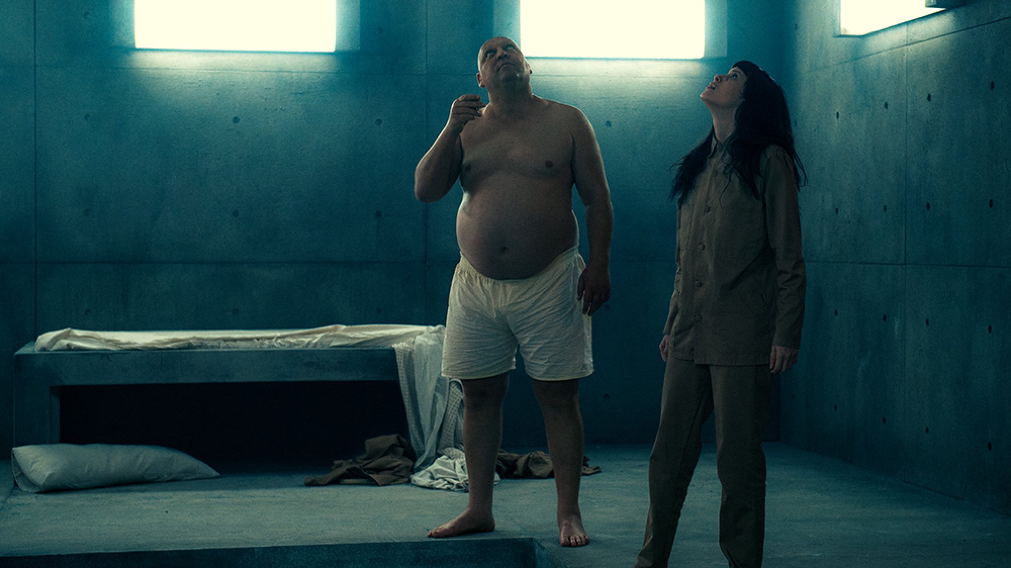 A shirtless man and a woman stand in a concrete room, looking up.