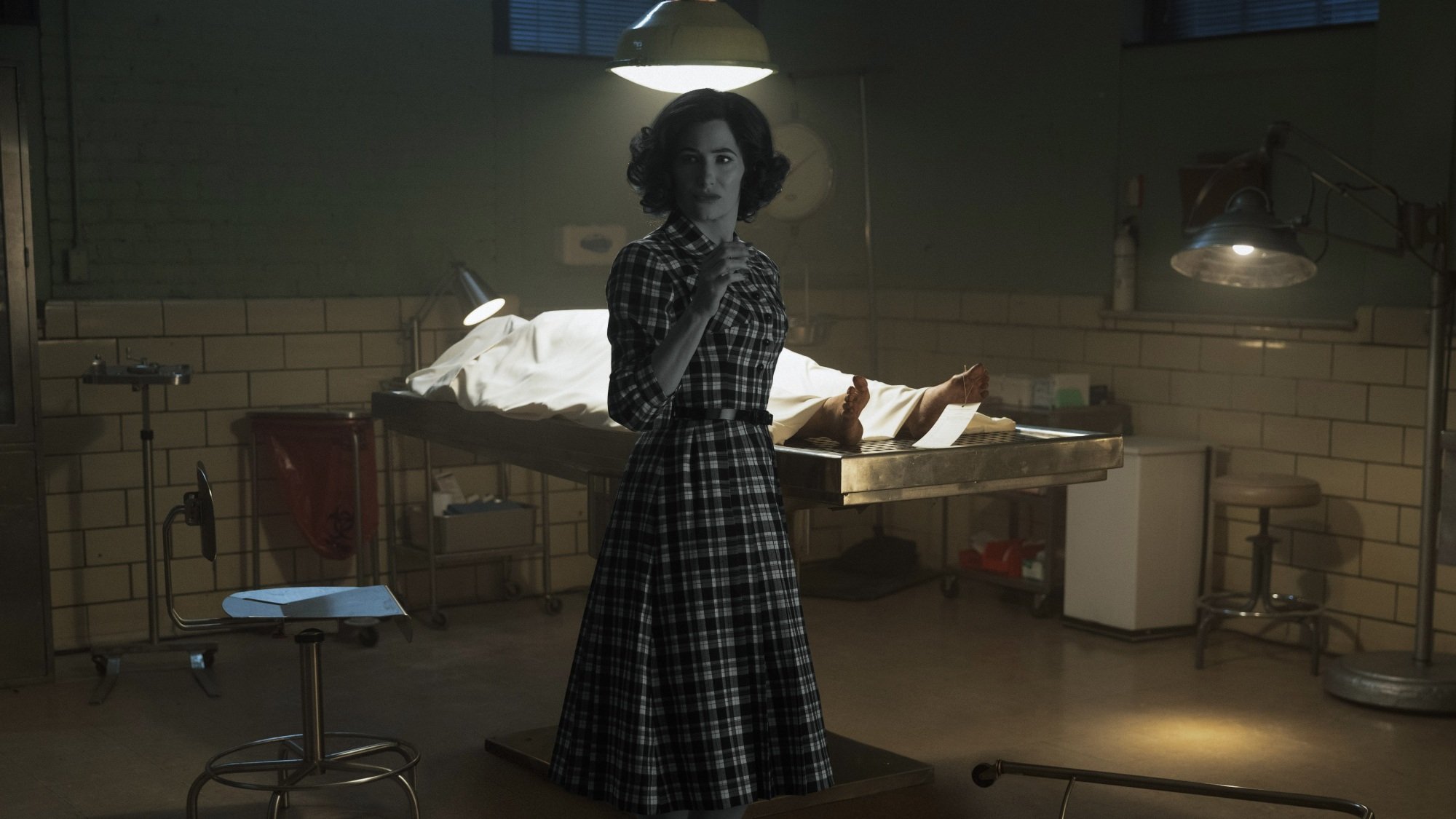 Agatha Harkness, in her black and white '50s sitcom outfit, stands in front of a dead body in a morgue.