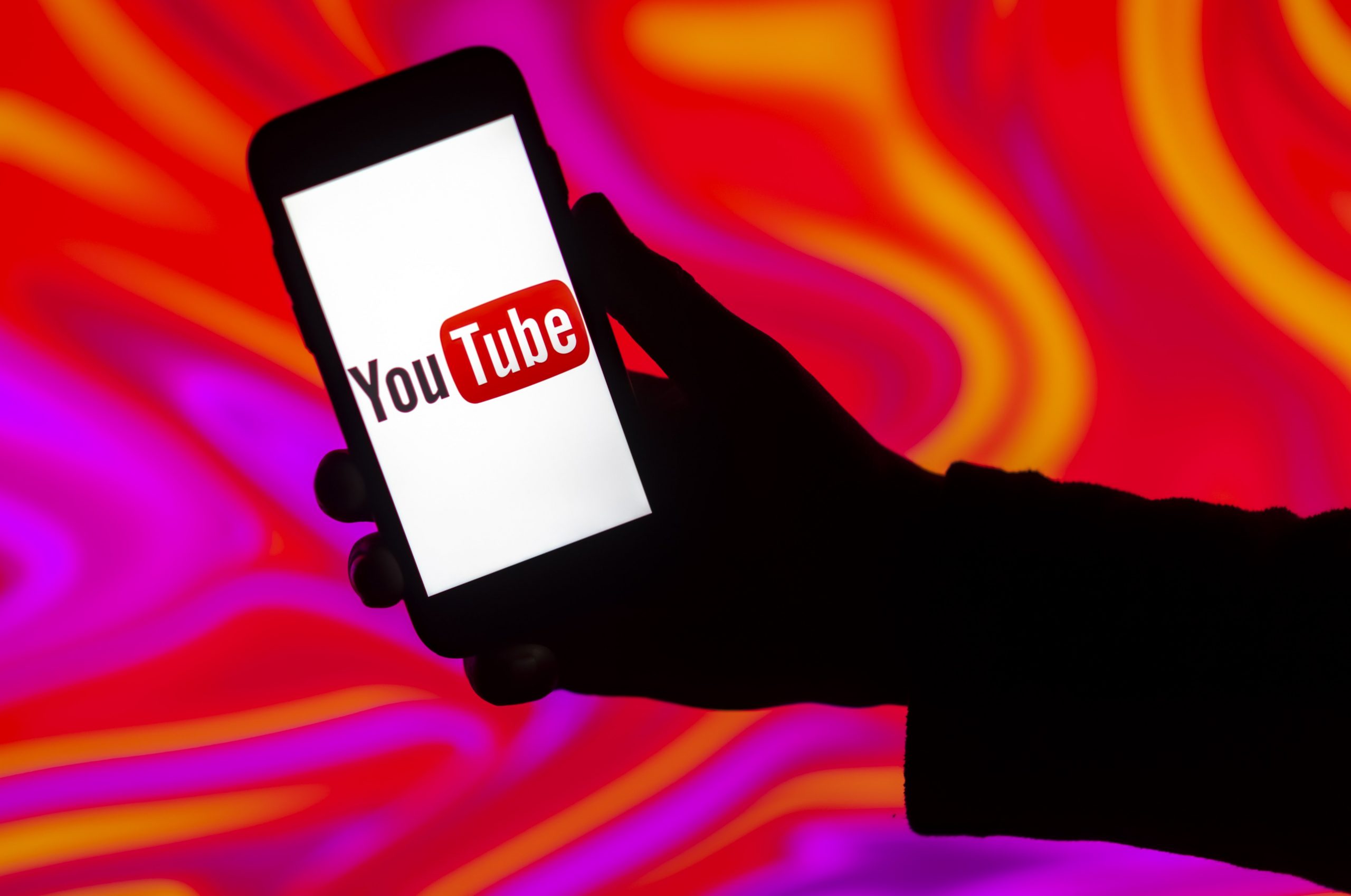 A silhouette of a person holding a phone displaying the YouTube logo surrounded by pink and orange tie dye. 