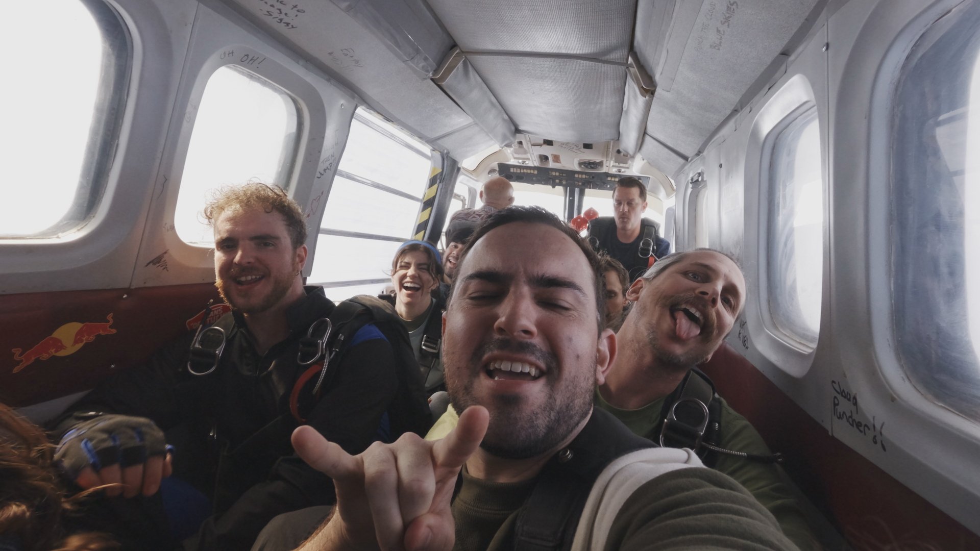 A group of skydivers in a plane grin at the camera.