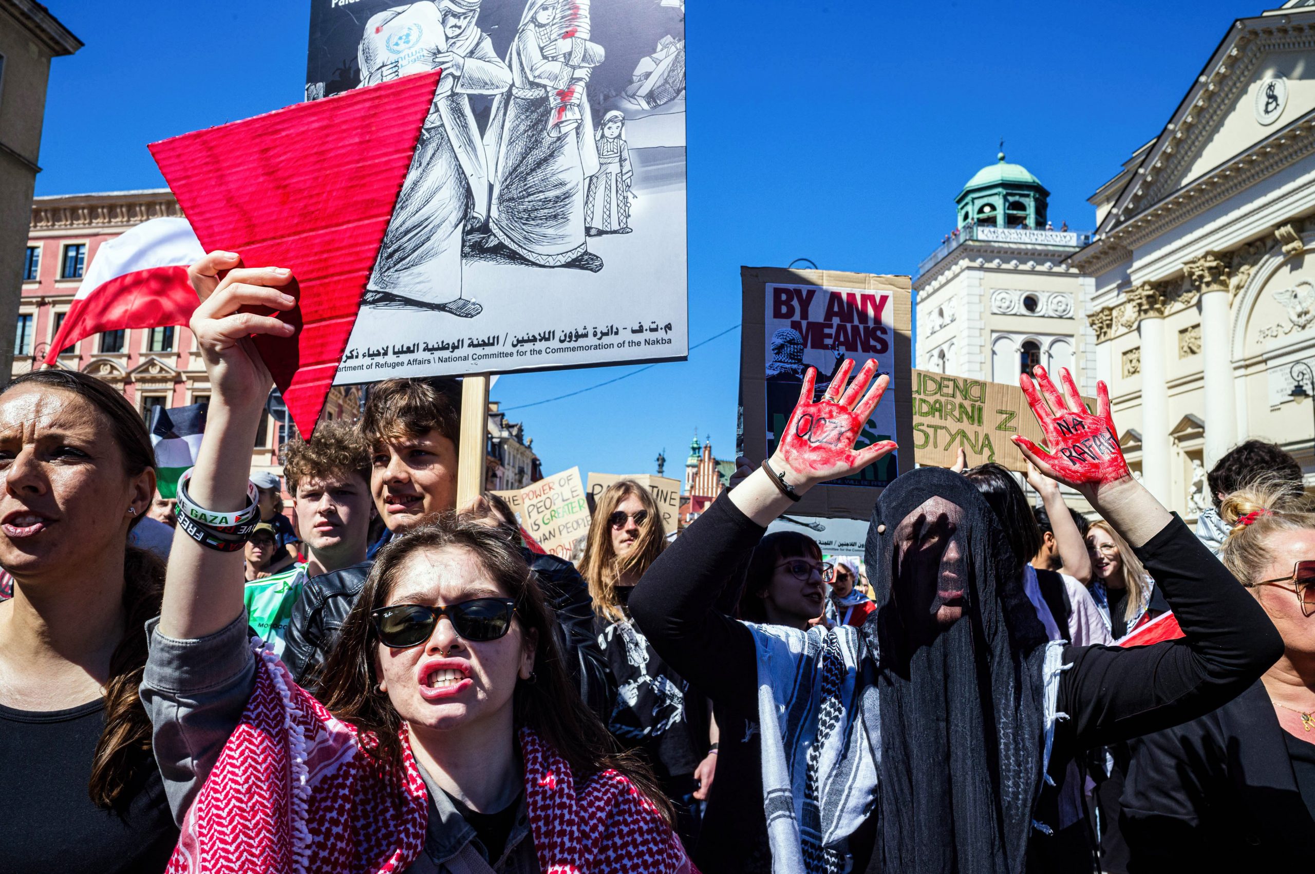 A group of pro-Palestinian protesters gather holding flags, signs, and large red triangles. 