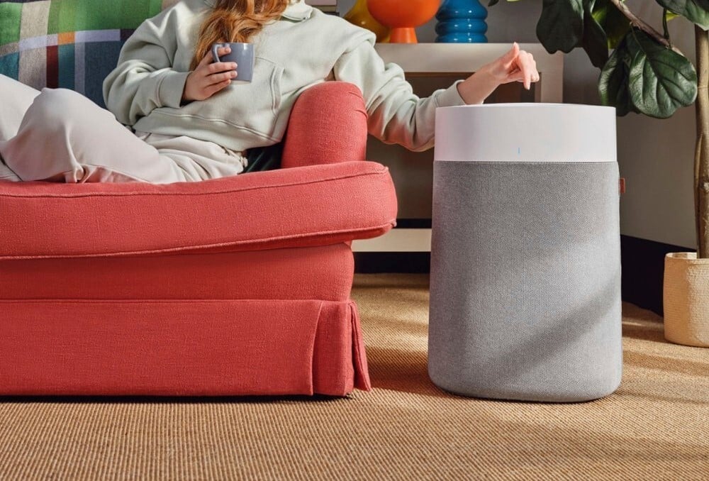 a blue air air purifier sits beside a woman sitting on a red couch