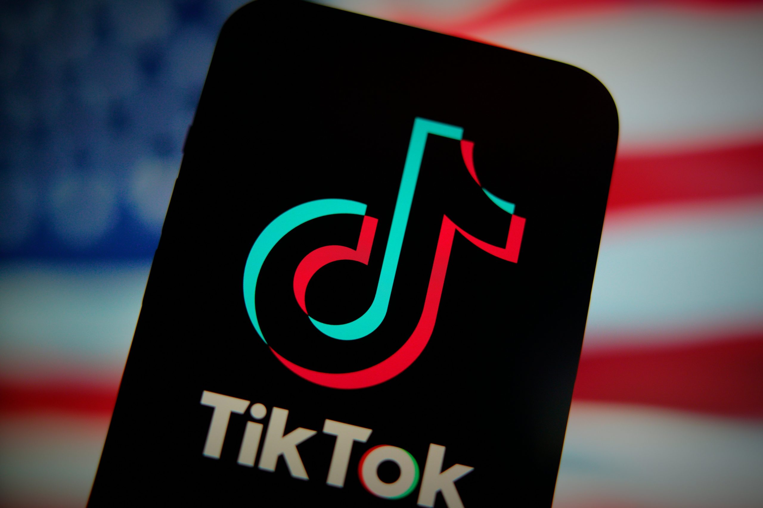 An phone displaying the TikTok logo in front of a blurry American flag. 