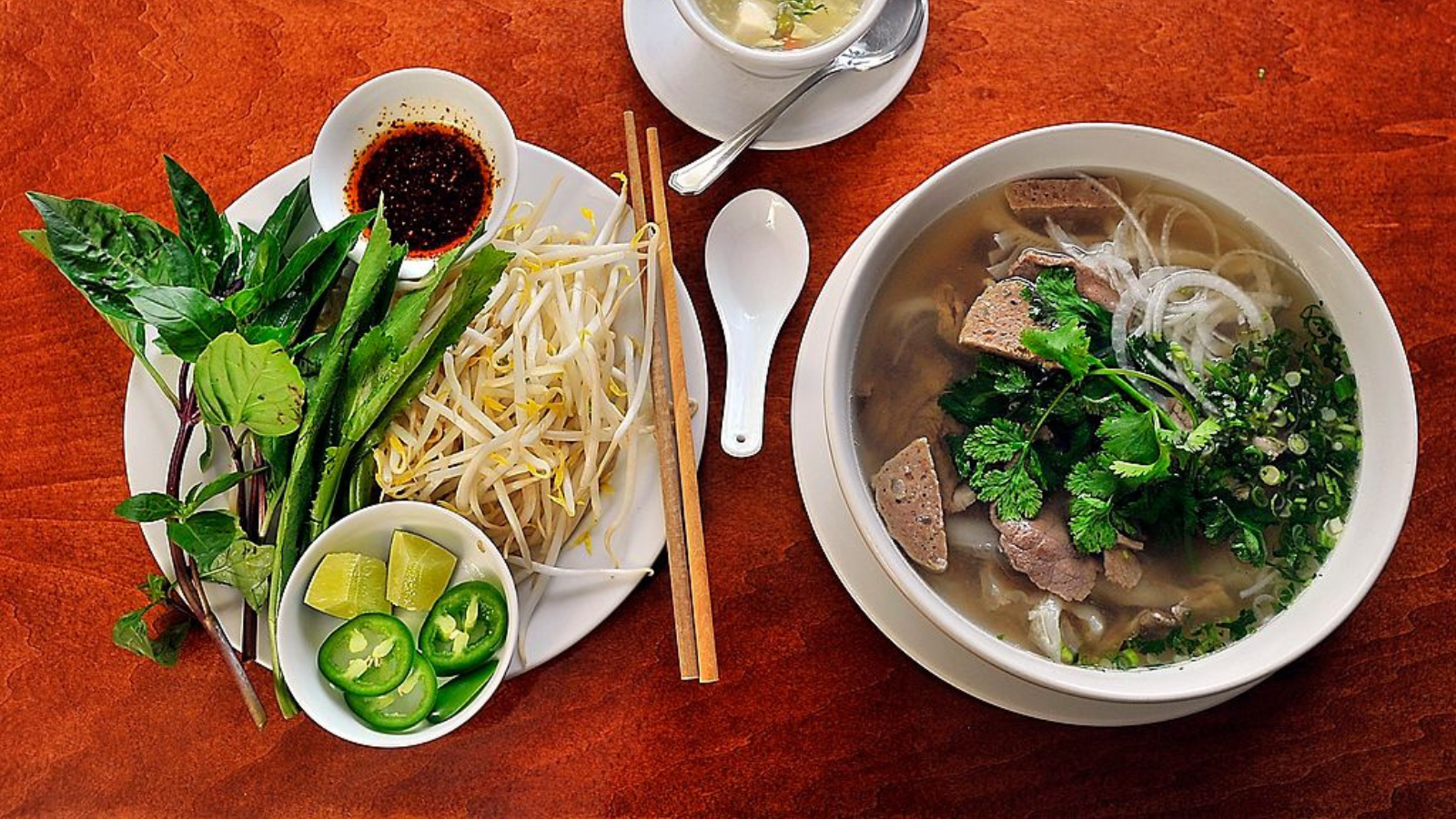 A bowl of beef pho (noodle soup) with a plate of fresh basil, bean sprouts, lime Jalapeno pepper slices, and hot oil. It comes with a small bowl of tofu soup, top, as a starter. In the Pho, right, are beef broth, noodles, onion, Asian cilantro, four kinds of meat: steak, brisket, beef tendon and meatball slices. 