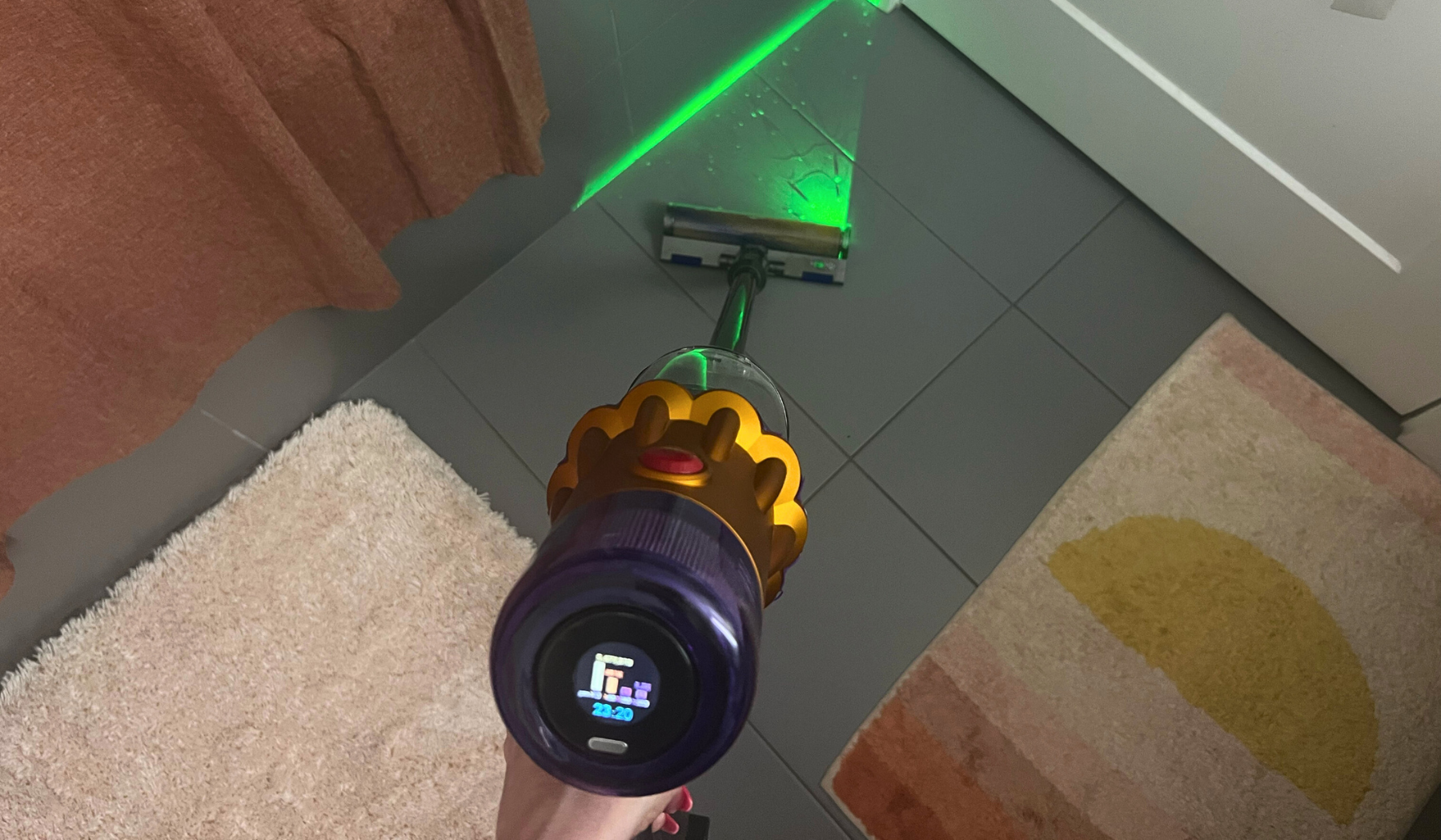 Hand cleaning tile bathroom floor with Dyson V12 Detect Slim vacuum and green laser highlighting debris on floor