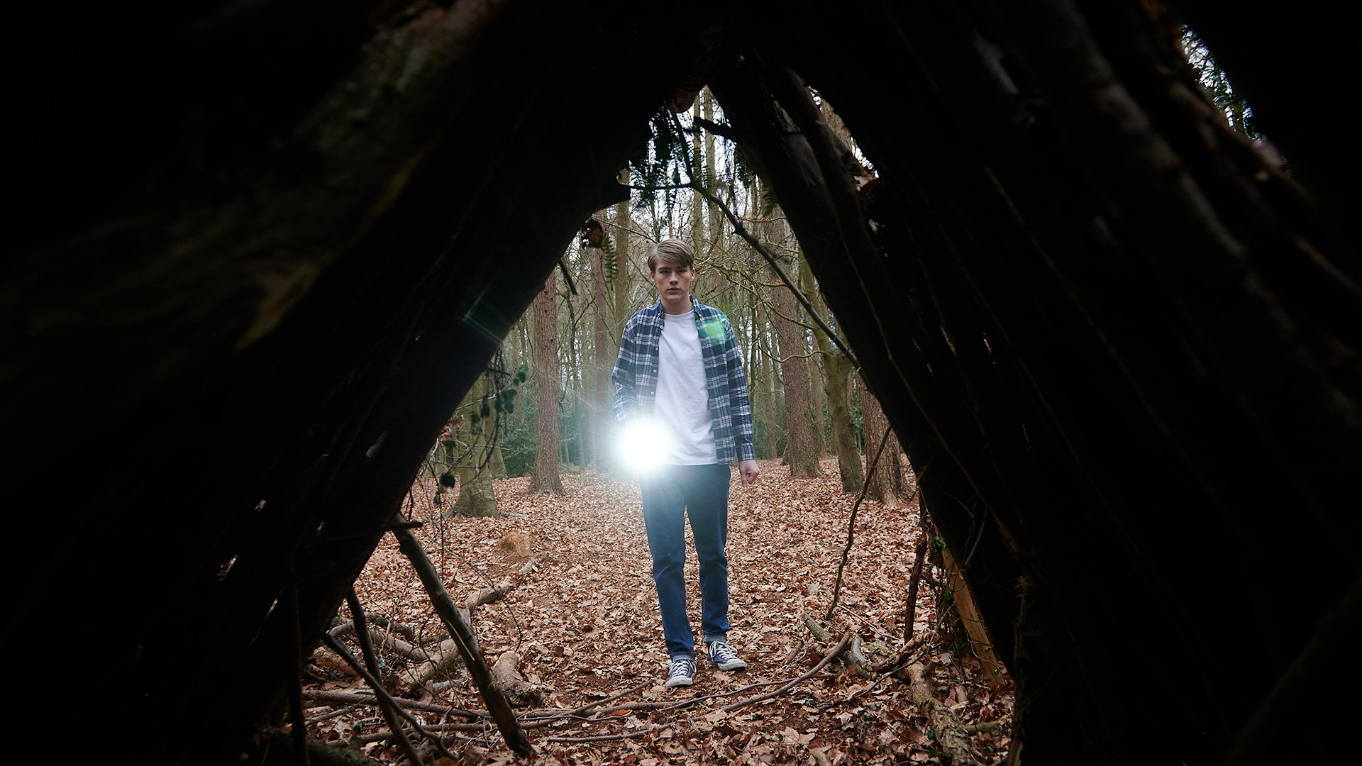 A boy approaches a wooden structure in the woods with a torch. He's viewed through the structure's triangular doorway.