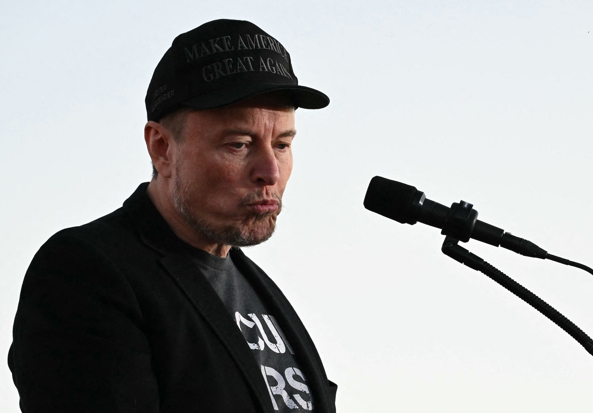 Elon Musk in his MAGA hat in front of a microphone.