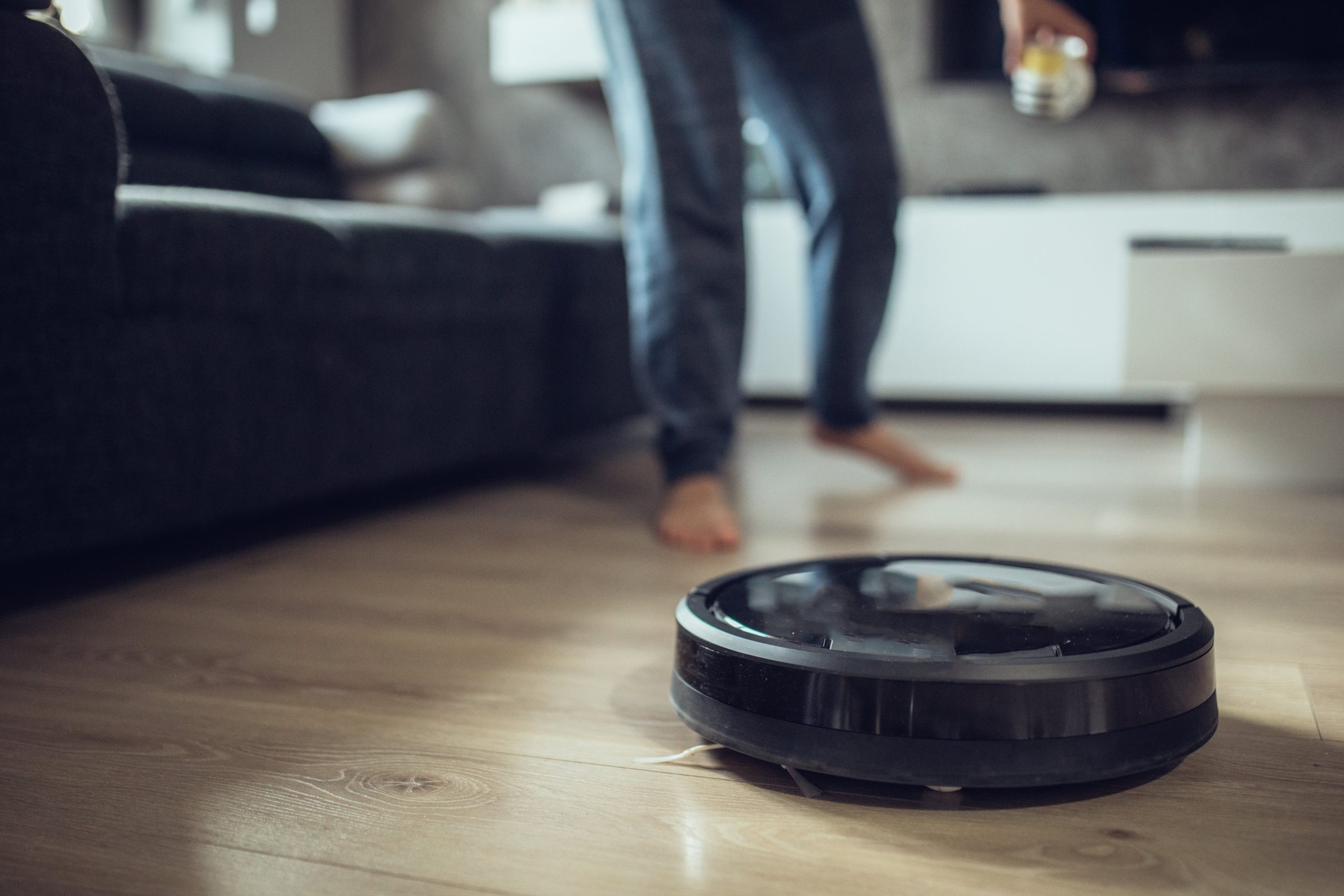 A robot vacuum on a wooden floor. A person's legs are seen running behind it.