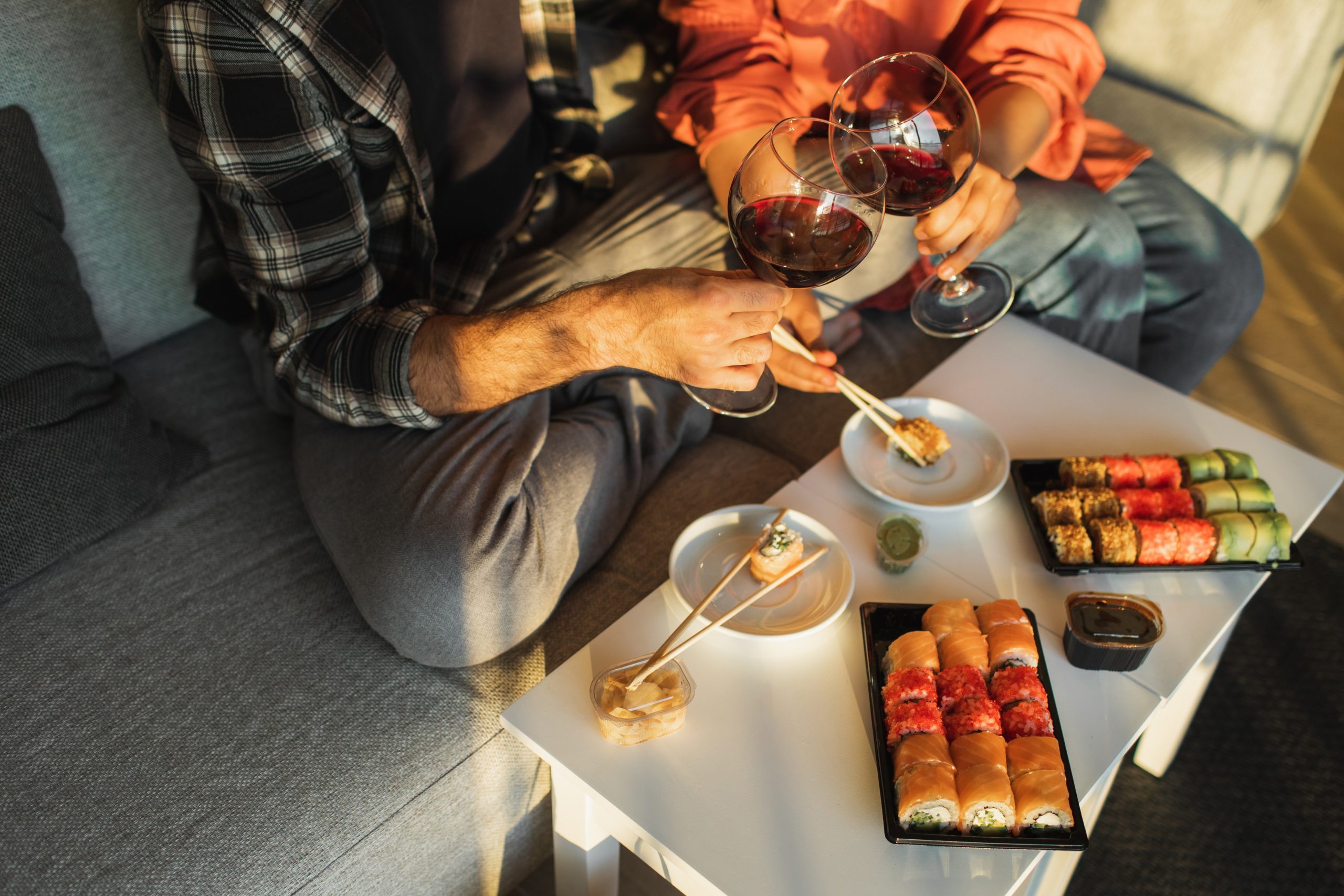 Two people sitting on couch holding wine glasses and eating sushi