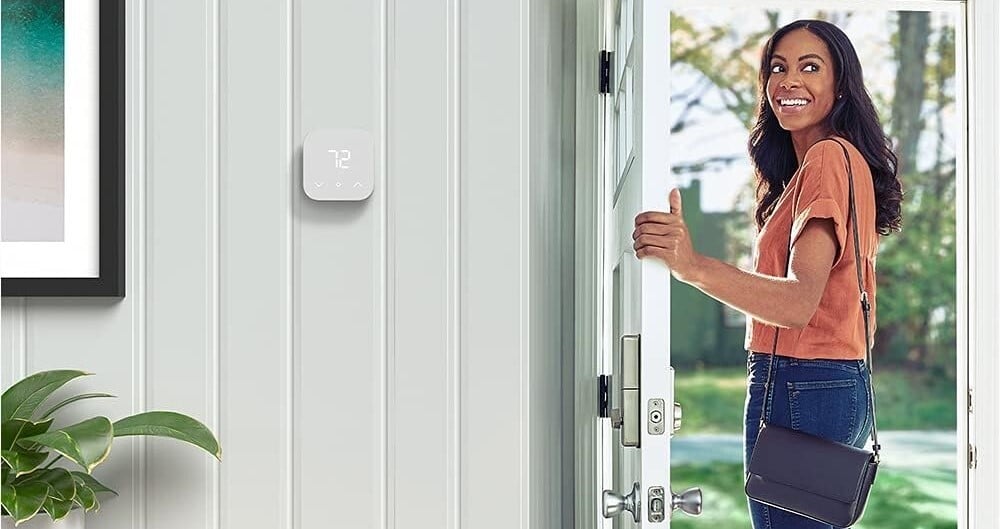 a person leaves through the front door of a home while looking back into the home and smiling. there's an amazon smart thermostat affixed to an interior wall inside the home