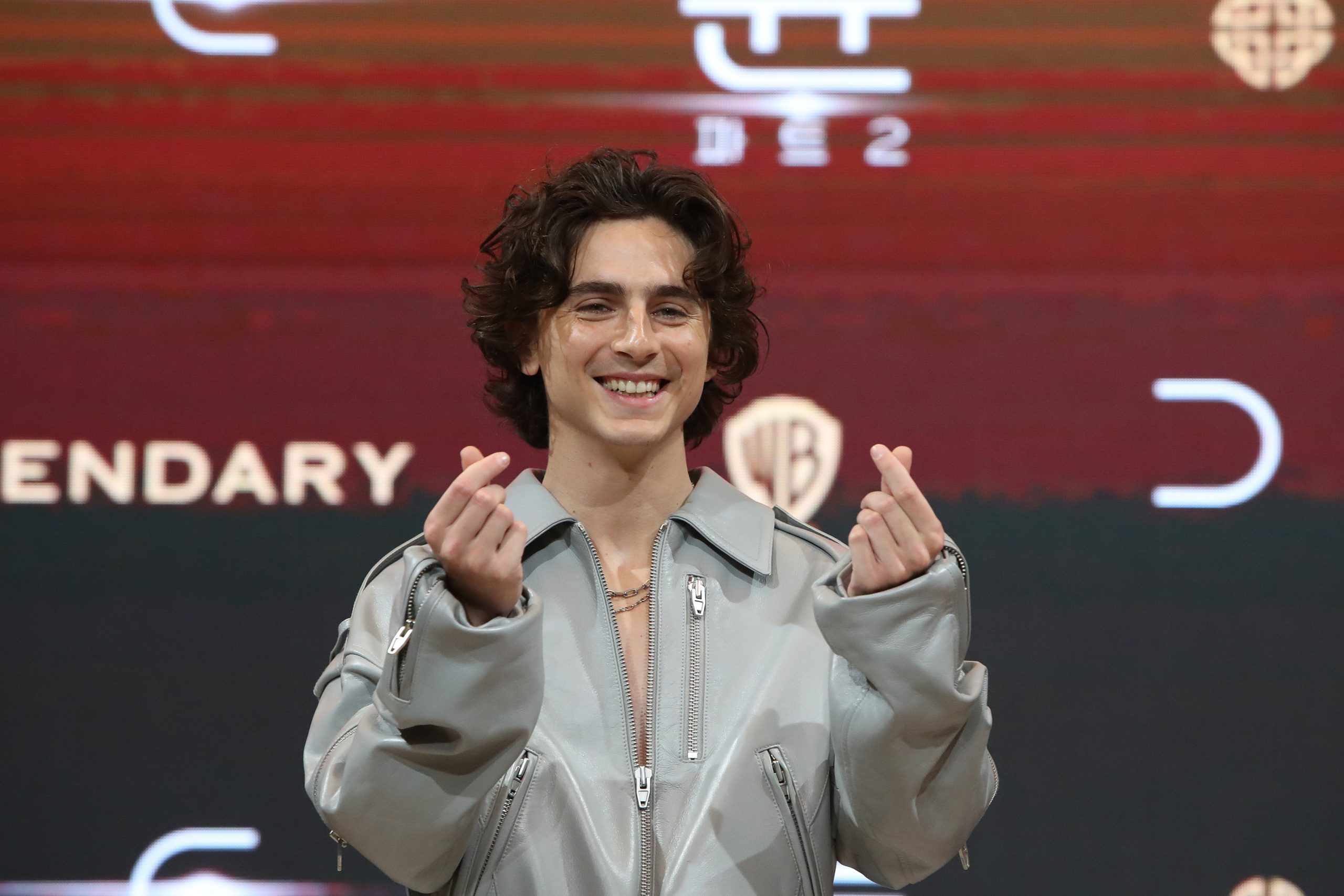 Timothée Chalamet smiling in a gray jumpsuit doing finger hearts.