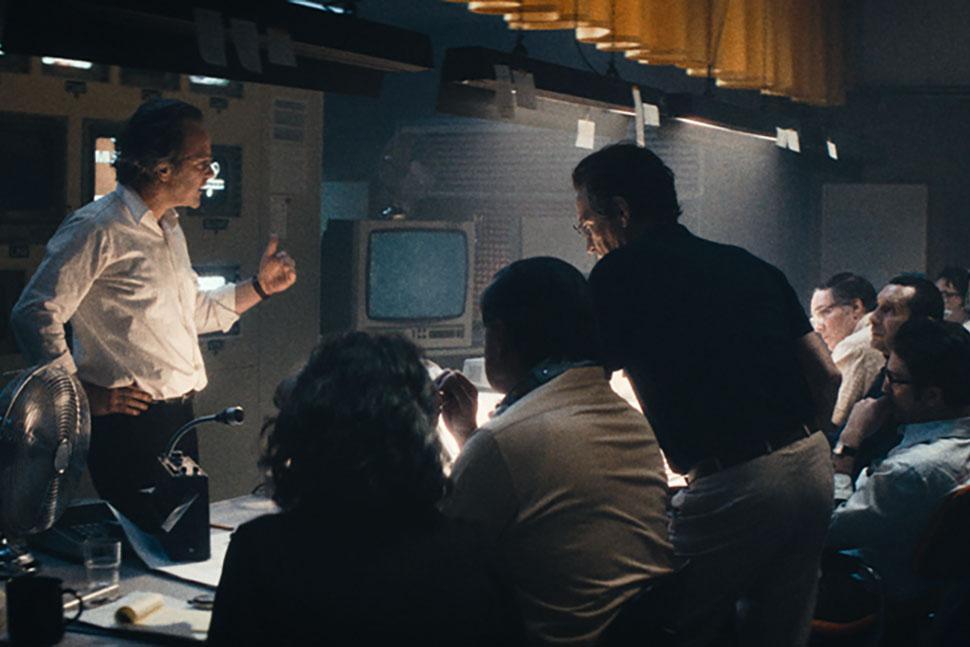 People sit and stand in a newsroom, talking and looking at one another. 