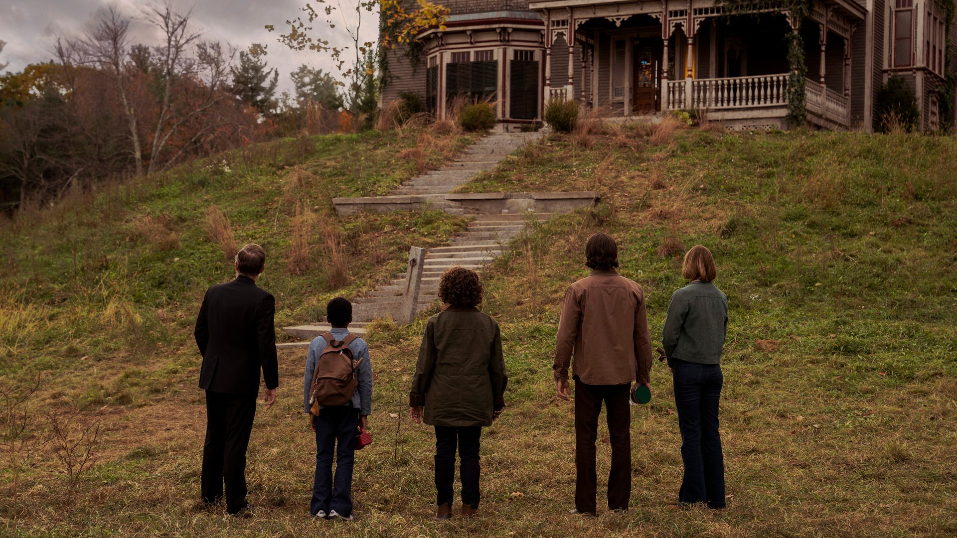 A group of people stand at the foot of a hill, looking up at a large house.