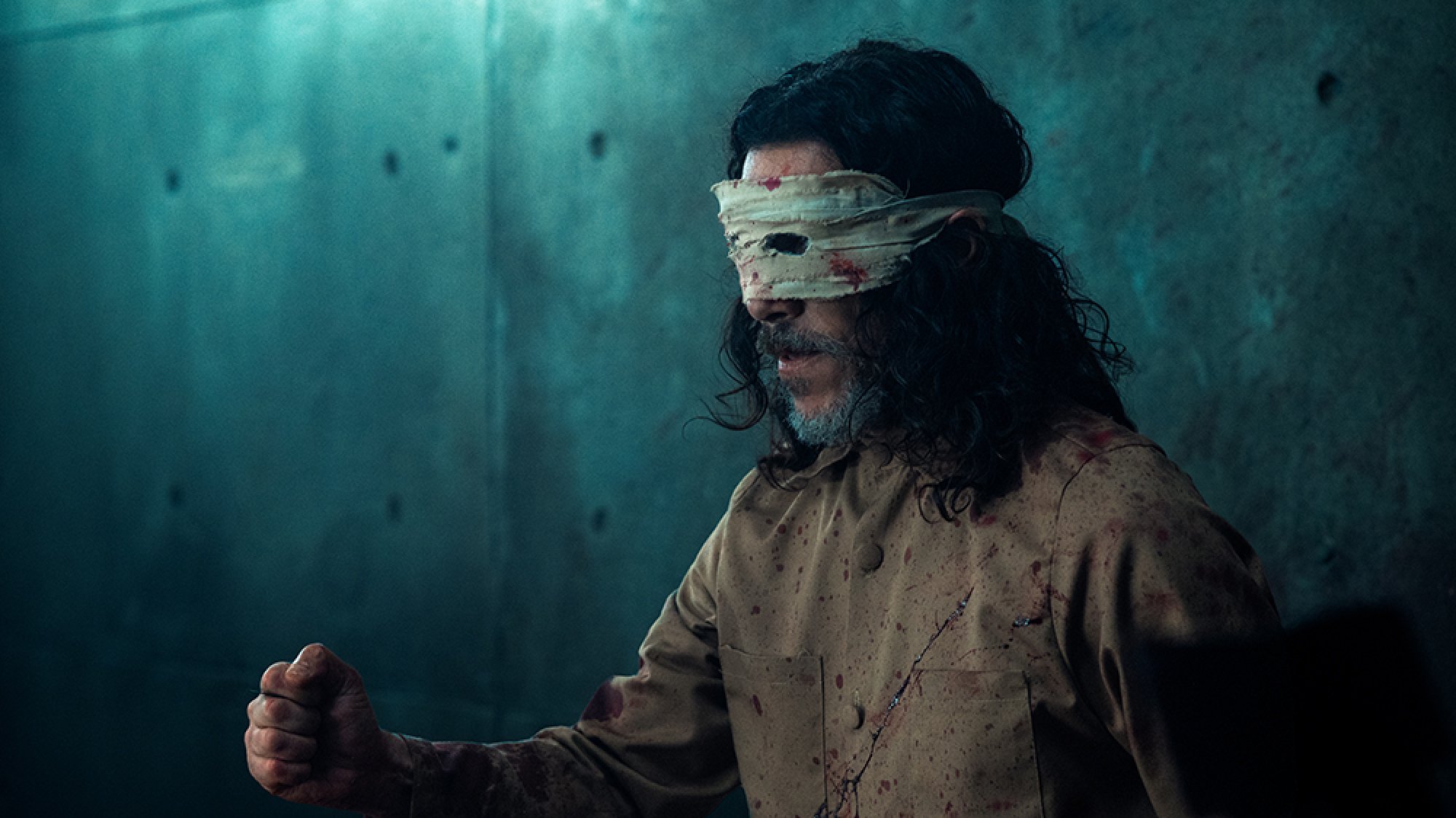 A man with bandages around his eyes stands in a concrete cell.