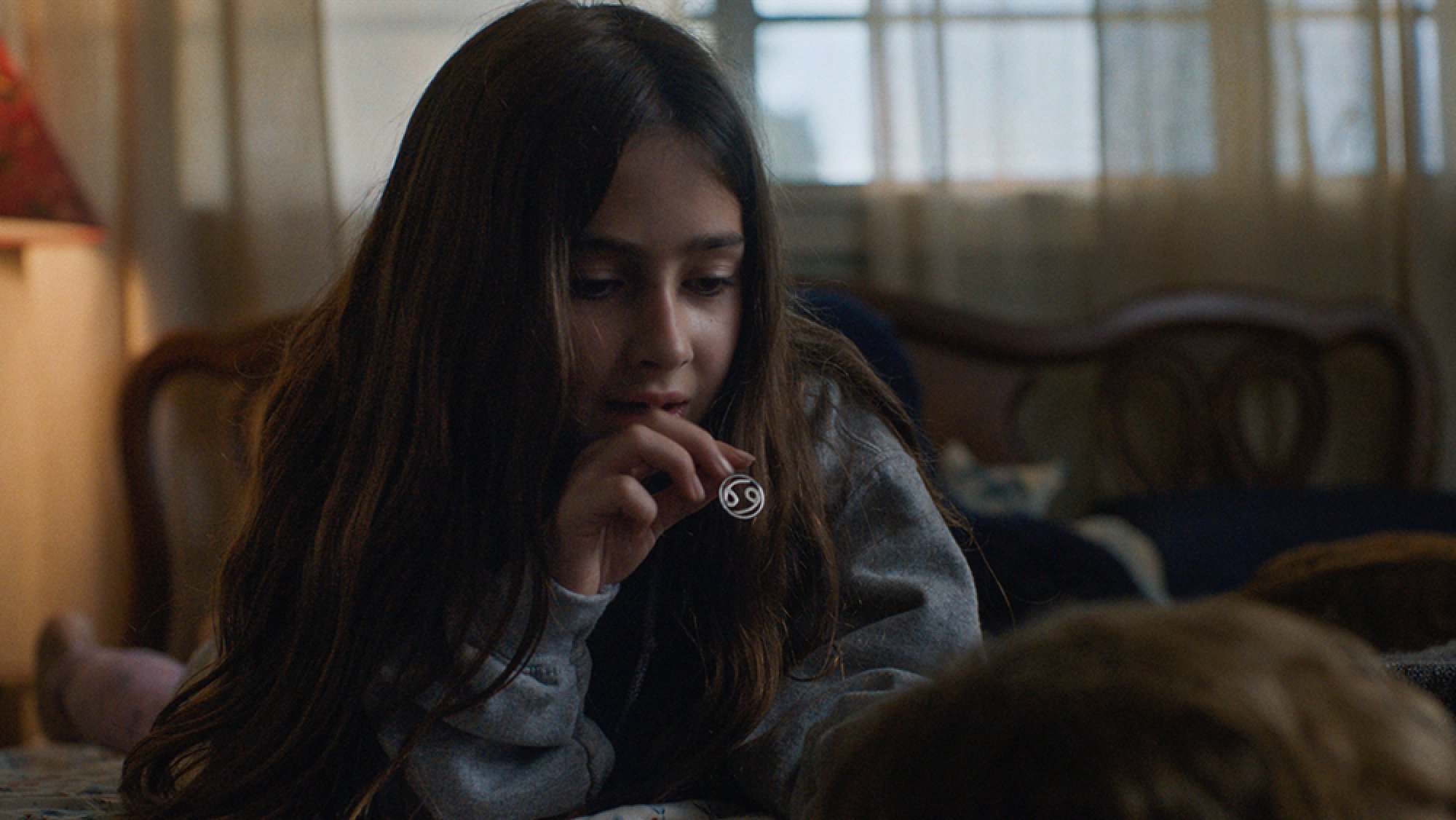 A little girl sits in a room holding her necklace.