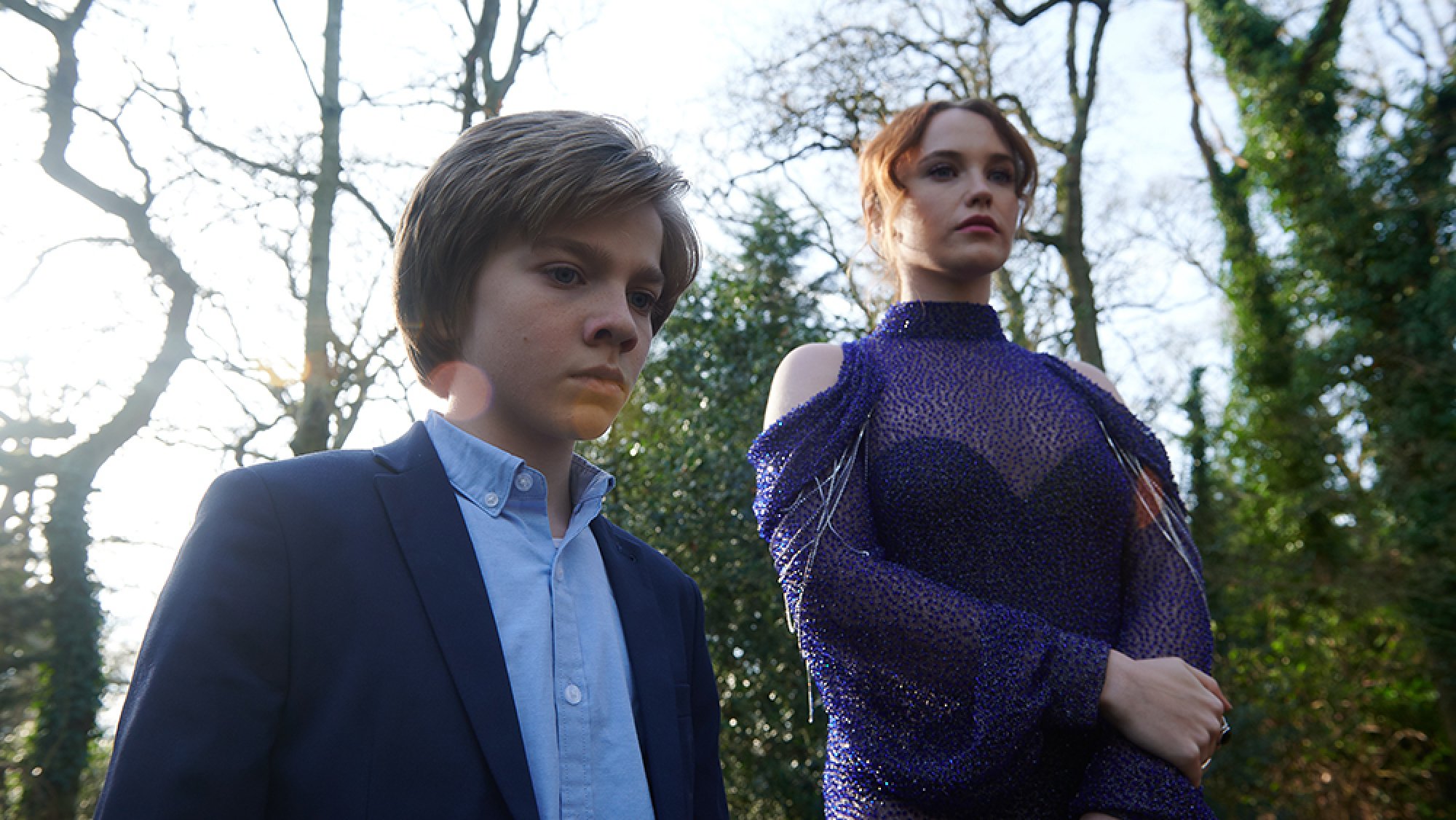 A young boy and a woman stand in smart clothing, looking sombre.