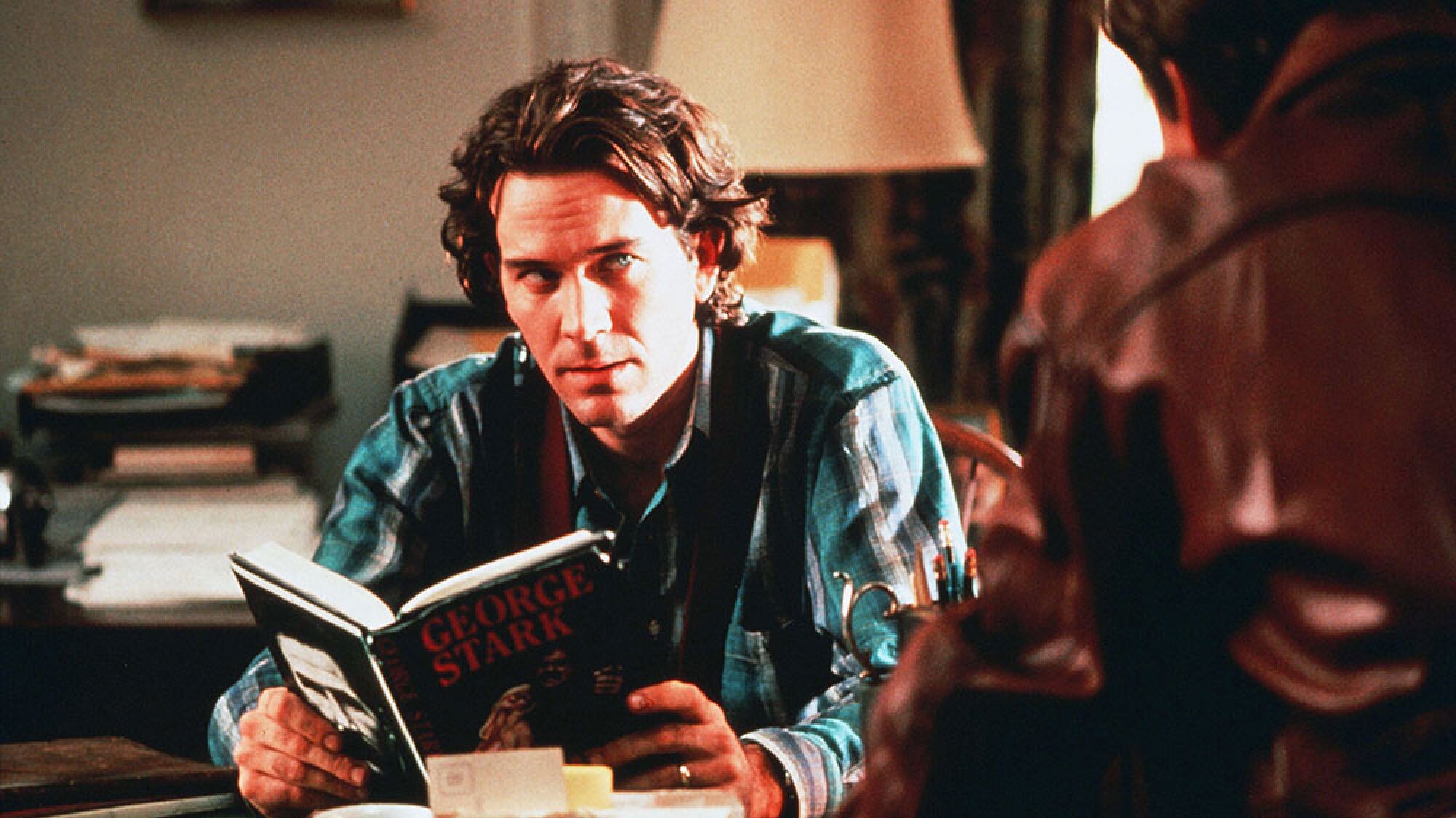 A man reads a book with the author's name "George Stark" on the cover.