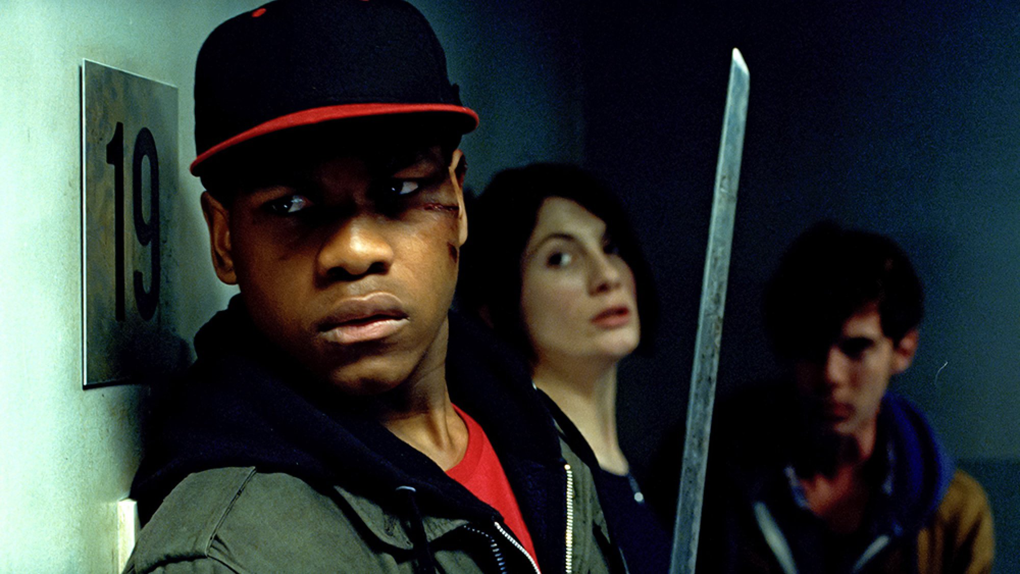 Three people stand in a corridor, looking scared. The man closest to the camera holds a sword and his face is cut.