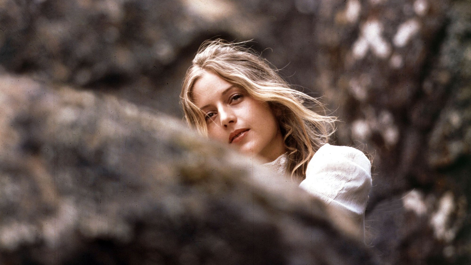 A young girl in a white dress sits between rocks in a still from "Picnic at Hanging Rock".