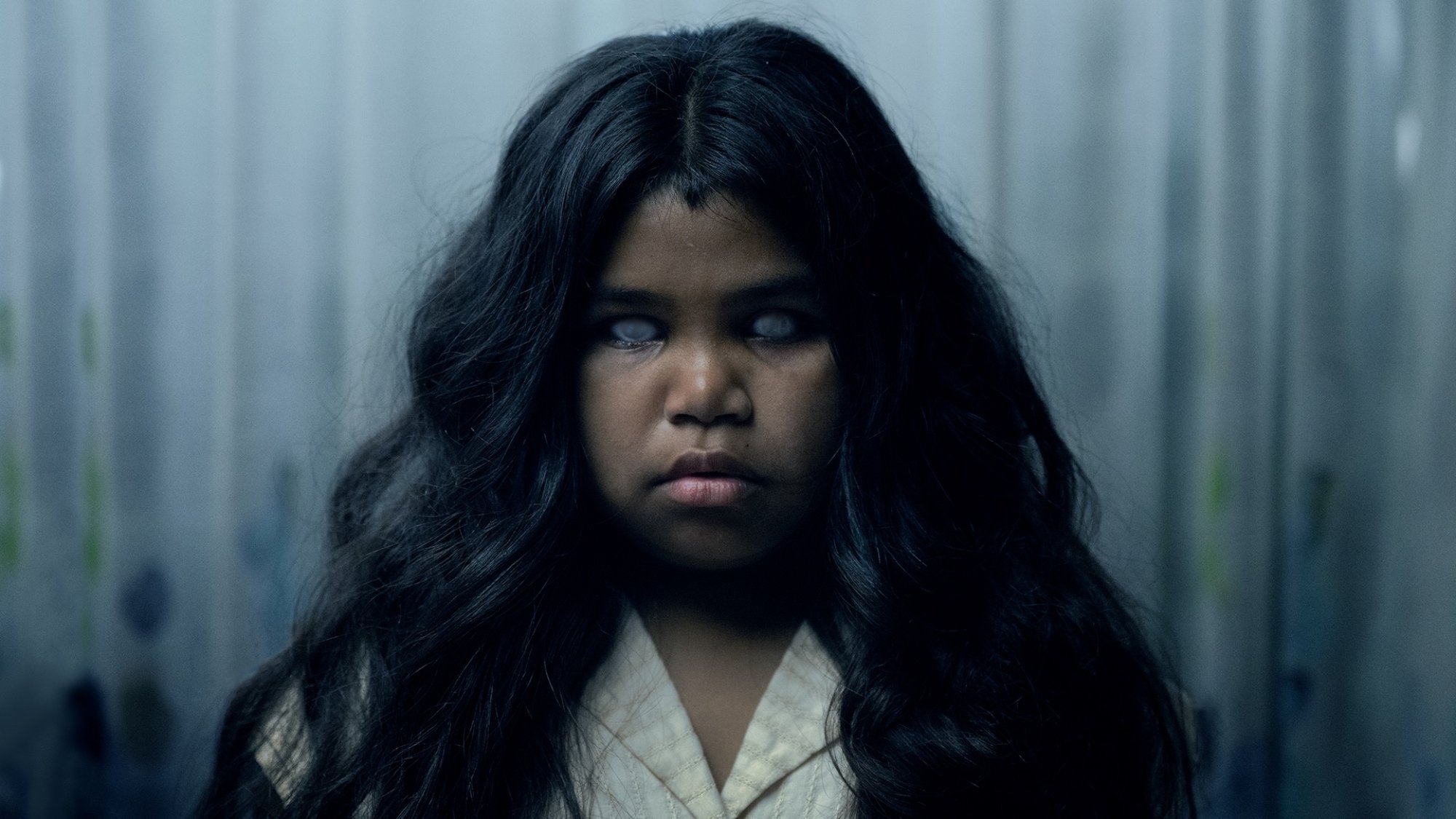 A young girl with white eyes stares at the camera in a still for "The Moogai".