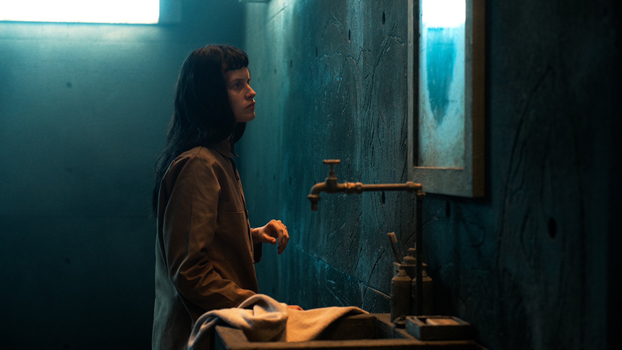 A woman stands in a dark concrete room, looking into a grimy mirror.