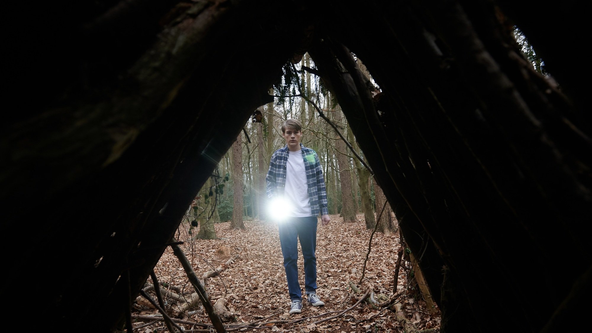 A young man shines a flashlight into a wooden structure in the woods.