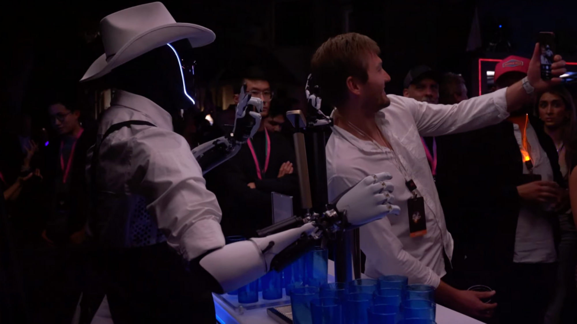 An Optimus robot bartender wearing a cowboy hat and showing a peace sign as a man takes a selfie with it.