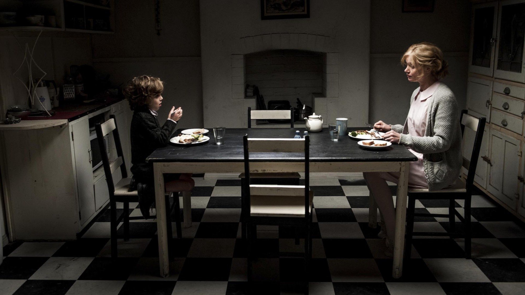 A child and a woman sit at opposite ends of a kitchen table in low lighting in "The Babadook"