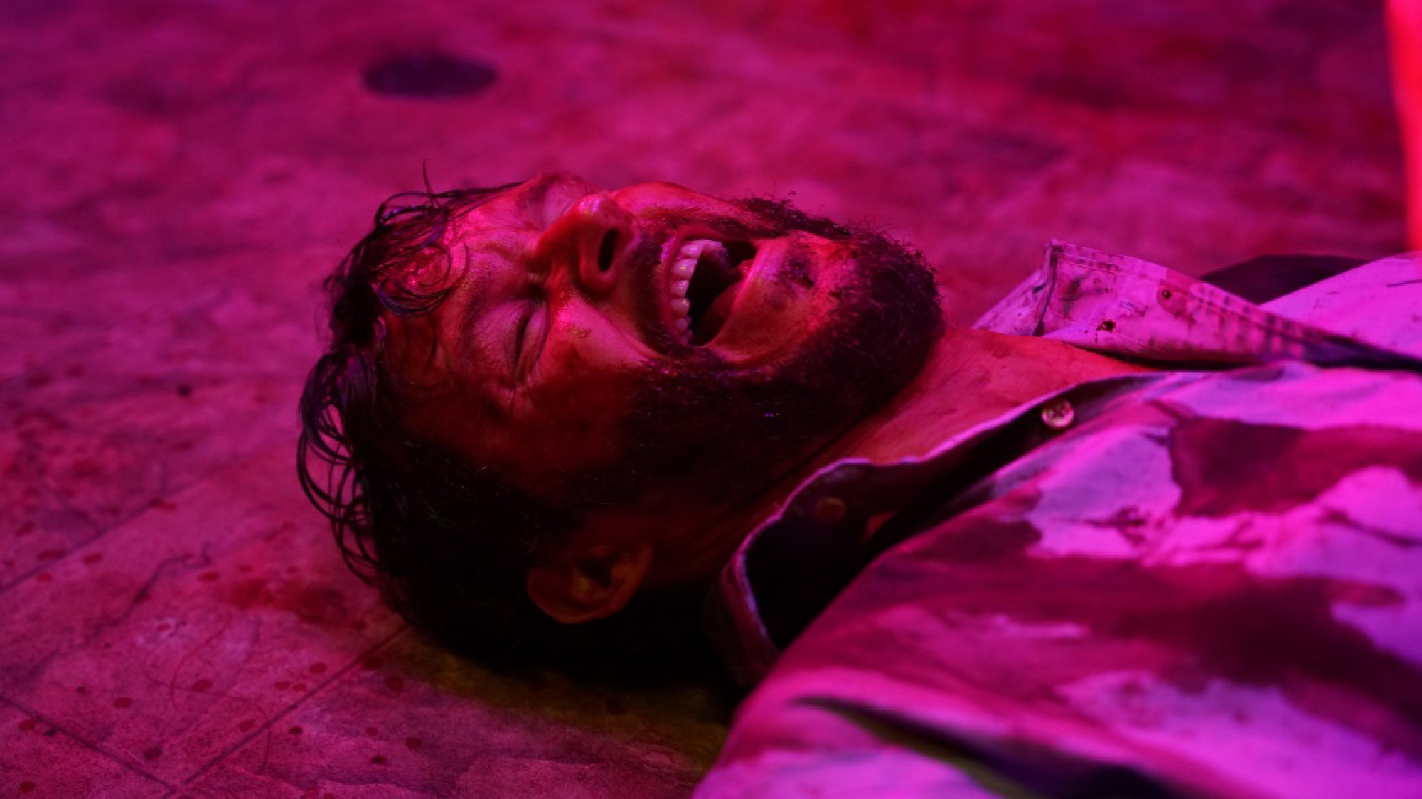 A screaming man lies on the floor of a public bathroom lit with purple light.