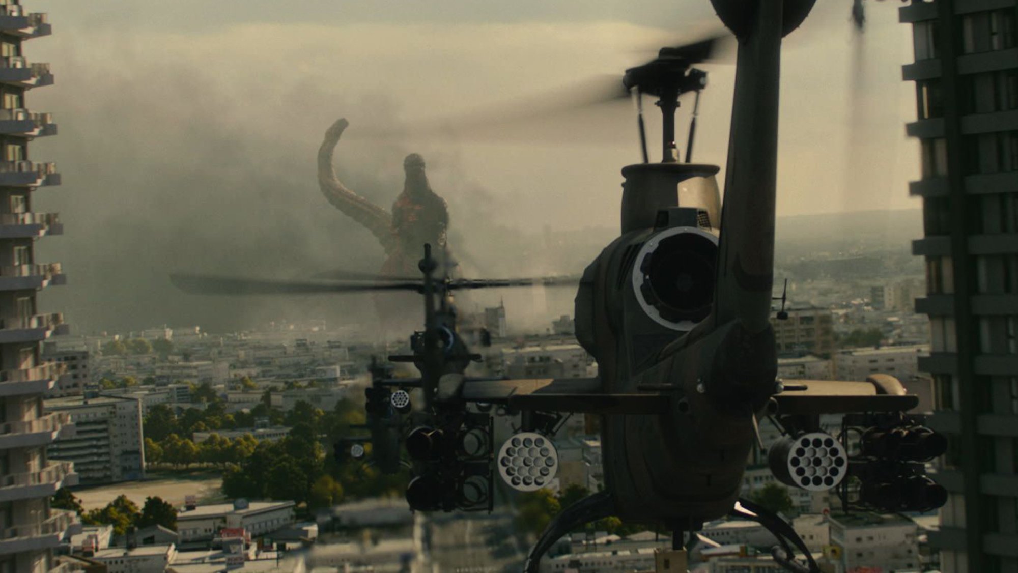 A helicopter hovers over a city as Godzilla looms on the horizon.