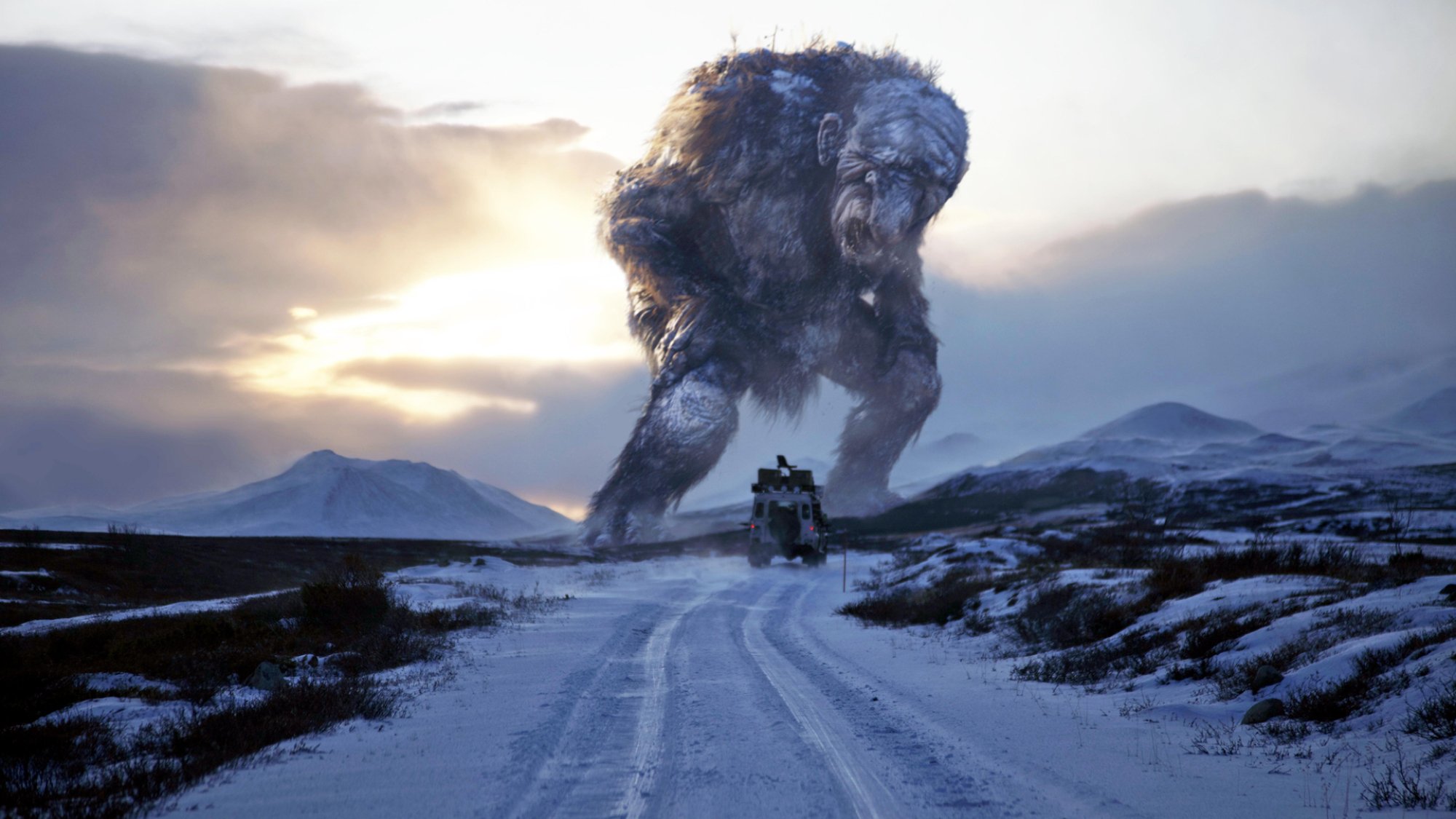 A giant troll stands astride a remote road in a wintry landscape, as a car drives toward it.