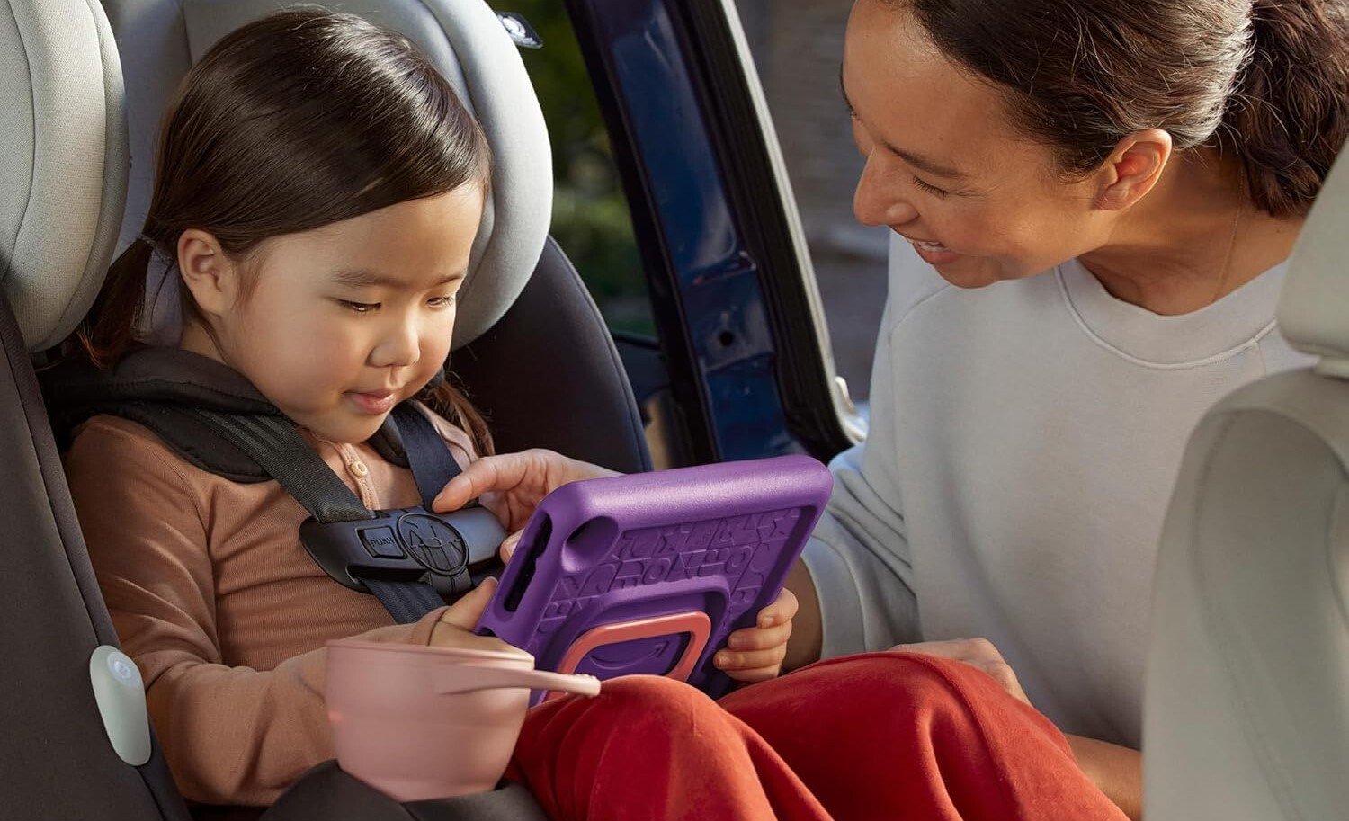 a small child uses an amazon kids tablet while sitting in the backseat of a car in a car seat while an adult clips the buckles of the car seat