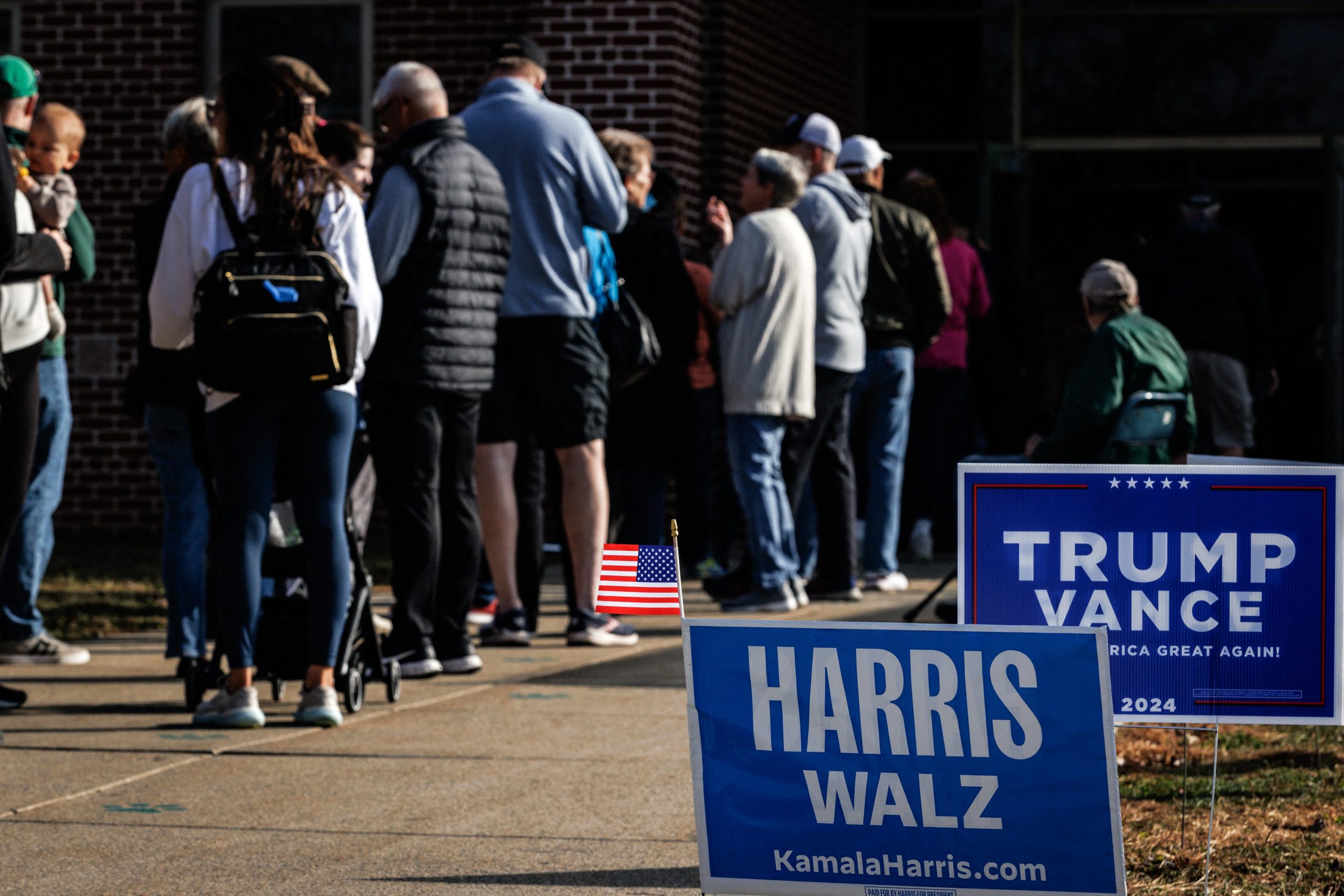 Harris and Trump yard signs are staked next to a line of voters entering a polling place.  