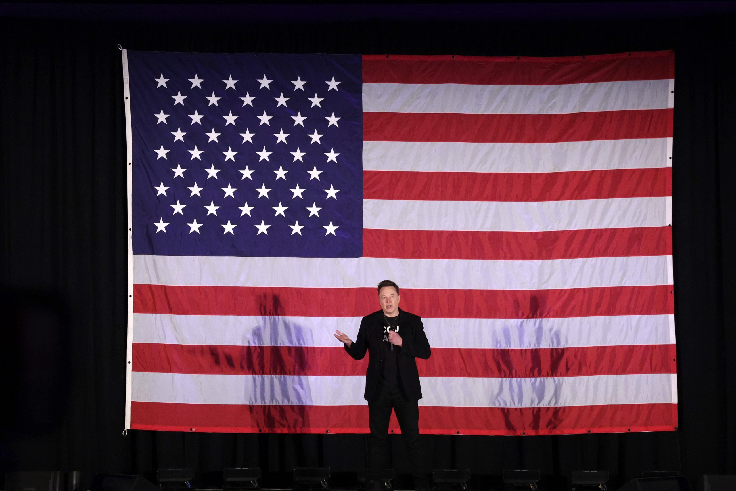 Elon Musk stands on stage in front of a giant American flag.