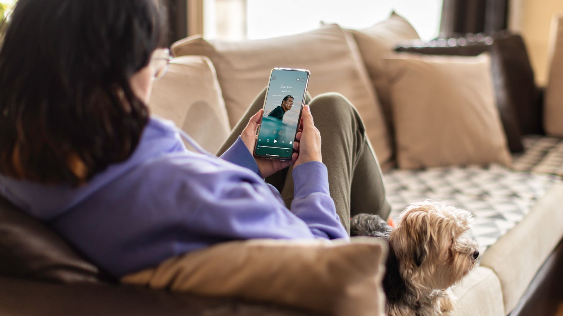 a person sits on a couch while looking at the calm app on a smartphone. A small dog is also lounging on the couch