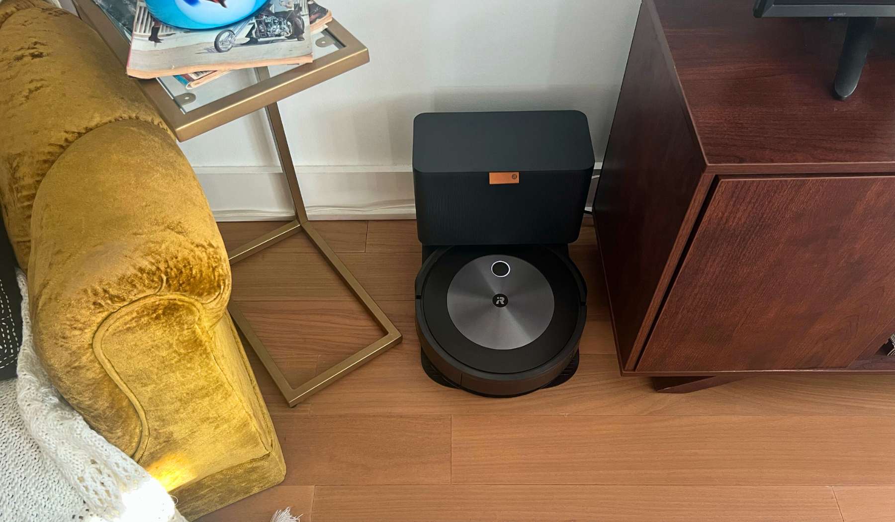 iRobot Roomba Combo j5+ and self-empty dock sitting against wall with yellow chair and TV stand in peripherals