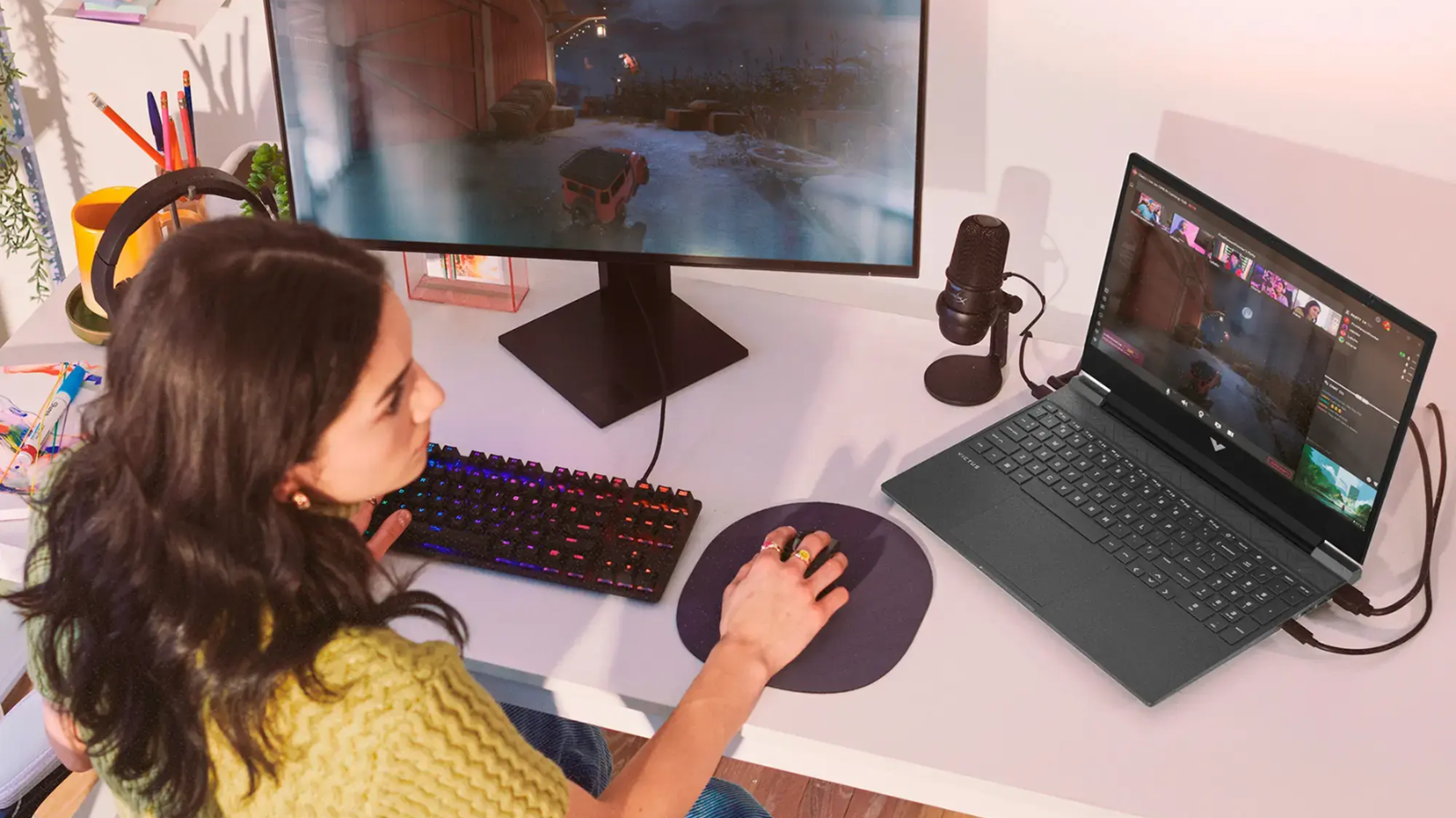 a top-down view of a woman playing games on a desktop computer that's hooked up to an hp victus 15 laptop