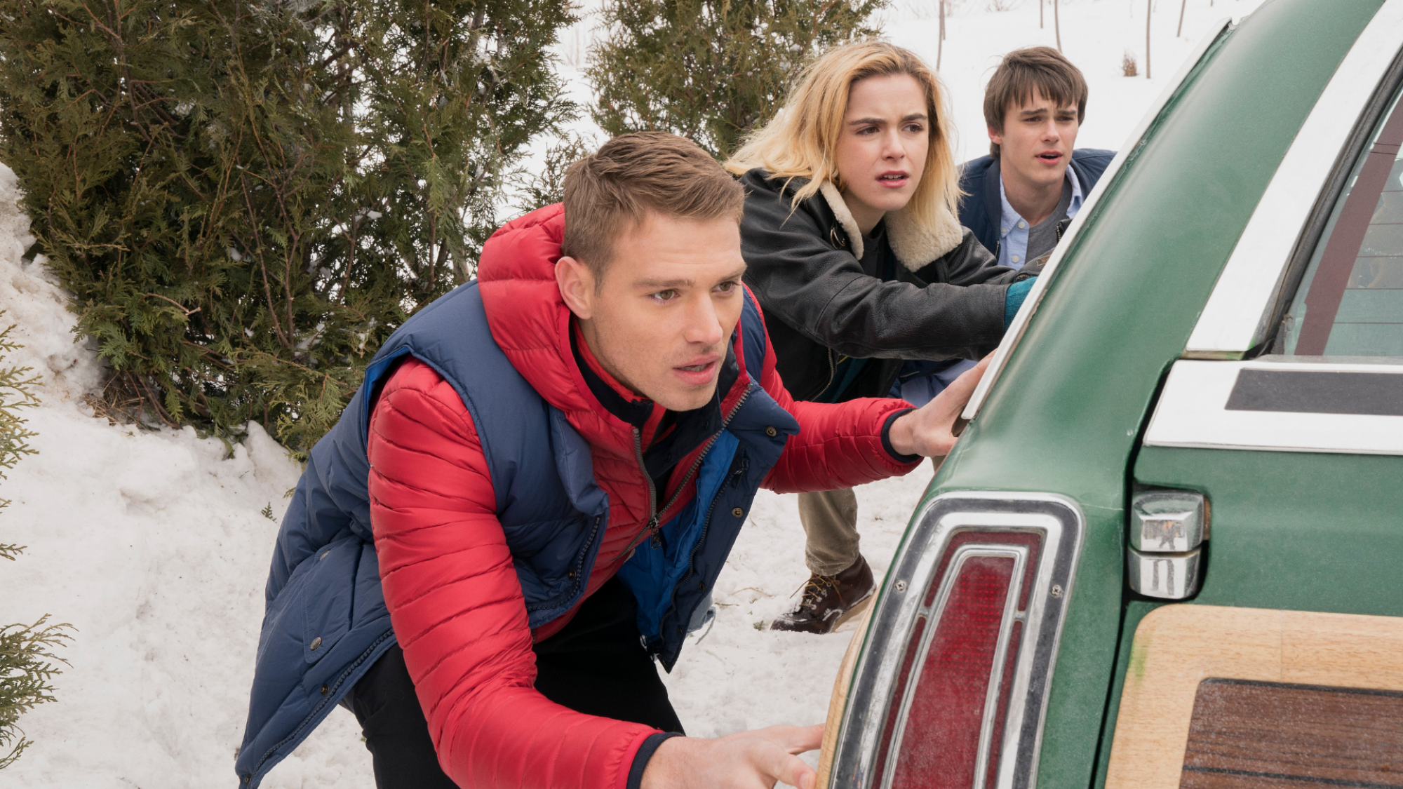 Teens hide behind a car in the snow.