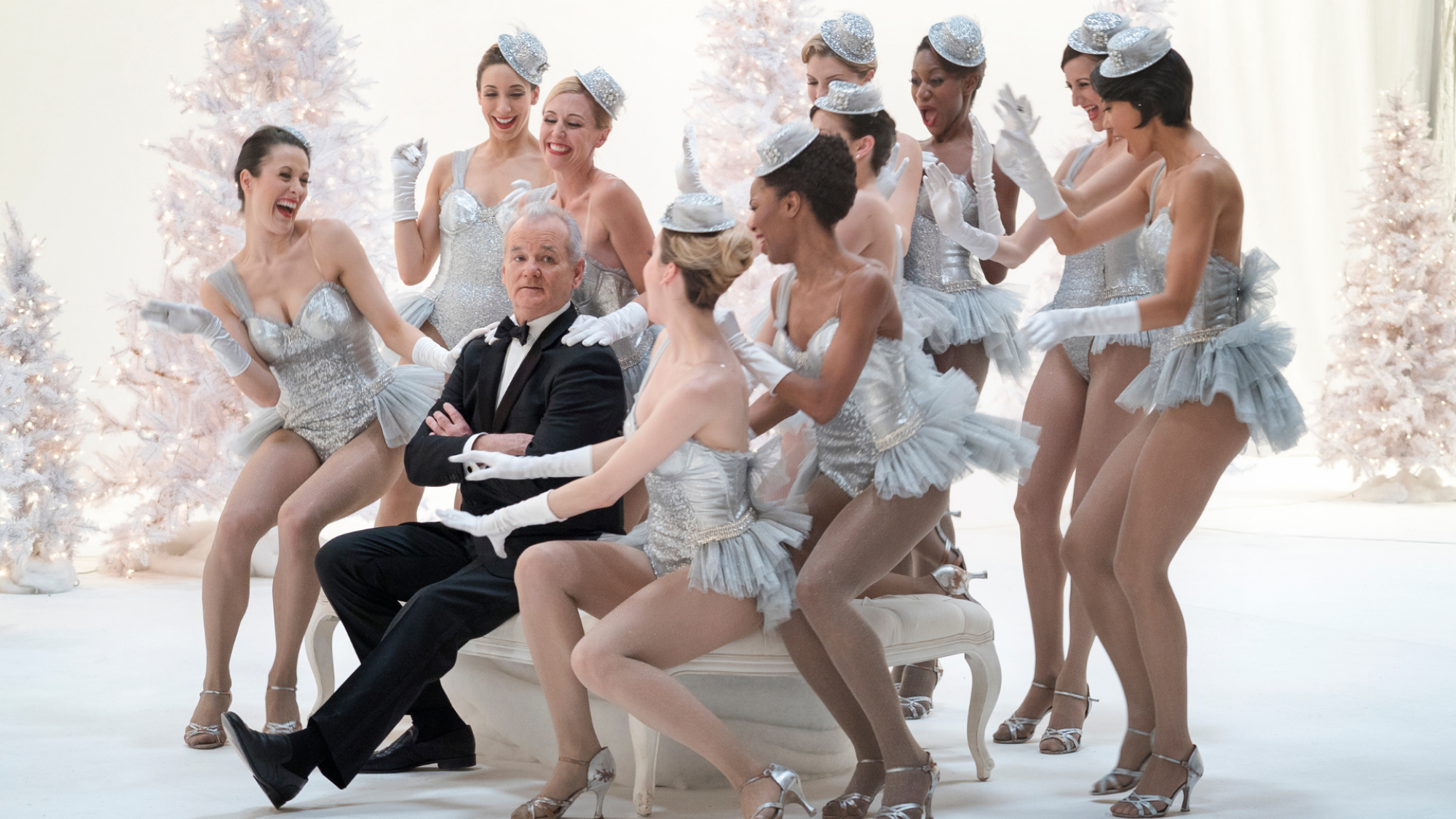 Dancers in sparkly costumes sit around Bill Murray, in a suit.